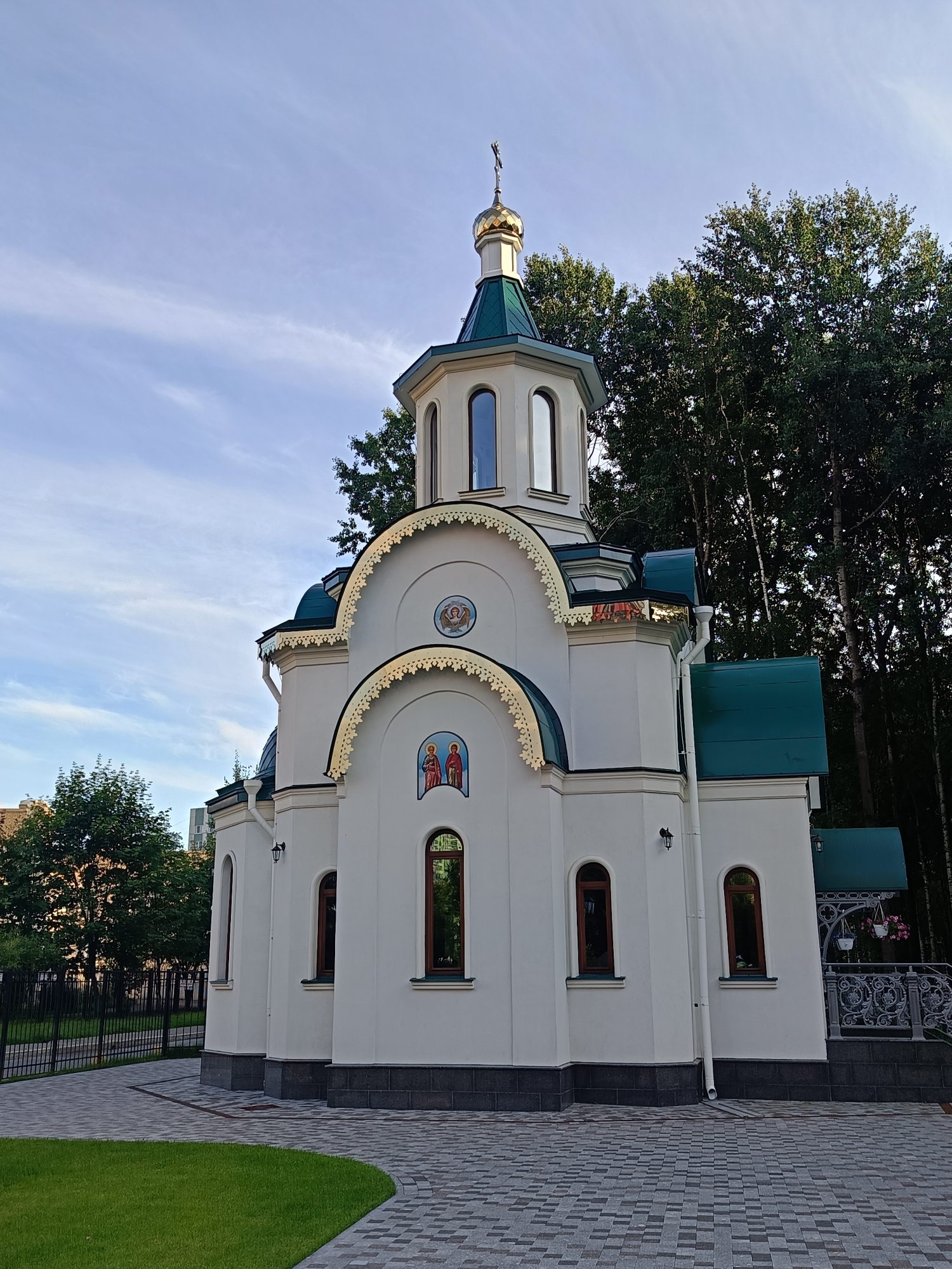 The image depicts a white orthodox church with a green roof and a golden dome topped with a cross. The church features arched windows and decorative golden trim. Two religious icons are visible on the facade. The church is surrounded by trees and a paved walkway. In the background, a black metal fence encloses a grassy area. The scene is bathed in natural light, suggesting it was taken during the daytime. The architectural style and religious iconography are characteristic of Russian Orthodox churches. Based on the architectural style and typical urban environment, this church is likely located in a residential or park area within the city of Saint Petersburg, Russia.