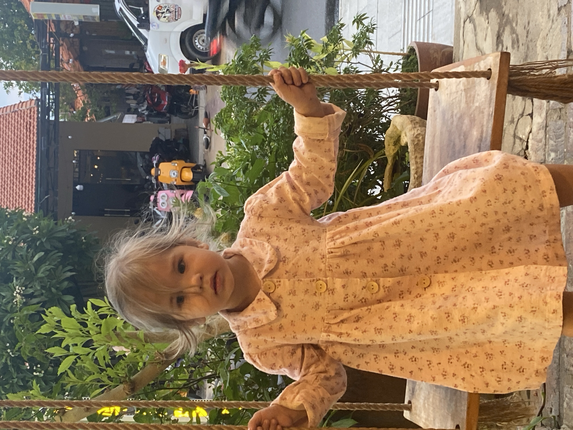A young girl in a floral dress sits on a wooden swing suspended by thick ropes. The swing is outdoors, in what appears to be a public area. The background shows a street scene in Phu Thuy Ward, Vietnam, featuring parked scooters and motorcycles, a white delivery van, and storefronts under an awning. Lush green foliage partially obscures the background elements. The girl looks toward the camera.