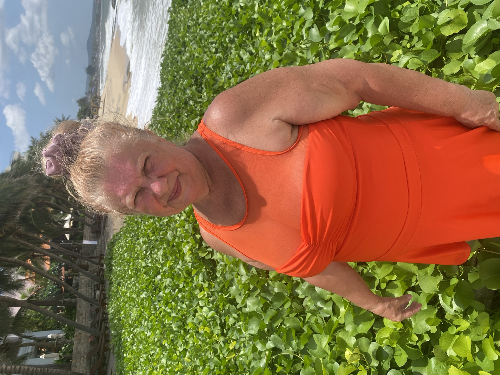 A woman with blonde hair in a bun, wearing an orange swimsuit, stands on a beach in Mũi Né, Vietnam. She is positioned in the foreground, surrounded by lush green vegetation. Behind her, a sandy beach meets the ocean with gentle waves. In the background, palm trees and buildings can be seen, suggesting a resort area. The sky is partly cloudy. The woman is looking towards the camera with a slight smile.