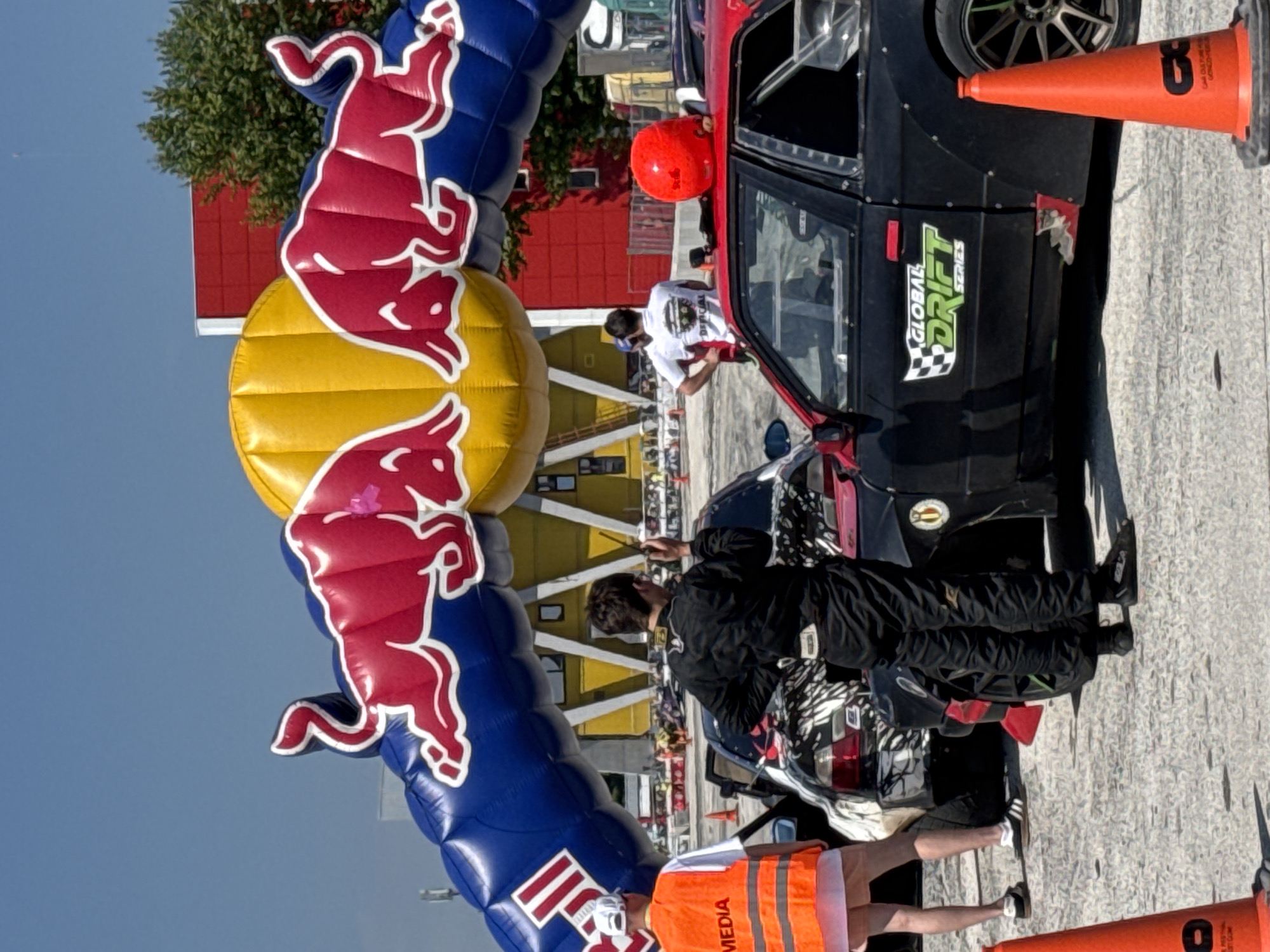 The image captures a scene at a drifting event, likely in Plovdiv, Bulgaria, given the presence of a Red Bull arch. A dark-colored drift car with "Global Drift Series" signage is parked near the arch. People in racing attire and media vests are present. A red helmet rests on the car’s roof. Orange traffic cones are placed around the car. The background features a yellow building with a structure reminiscent of stands. The sky is clear and blue.