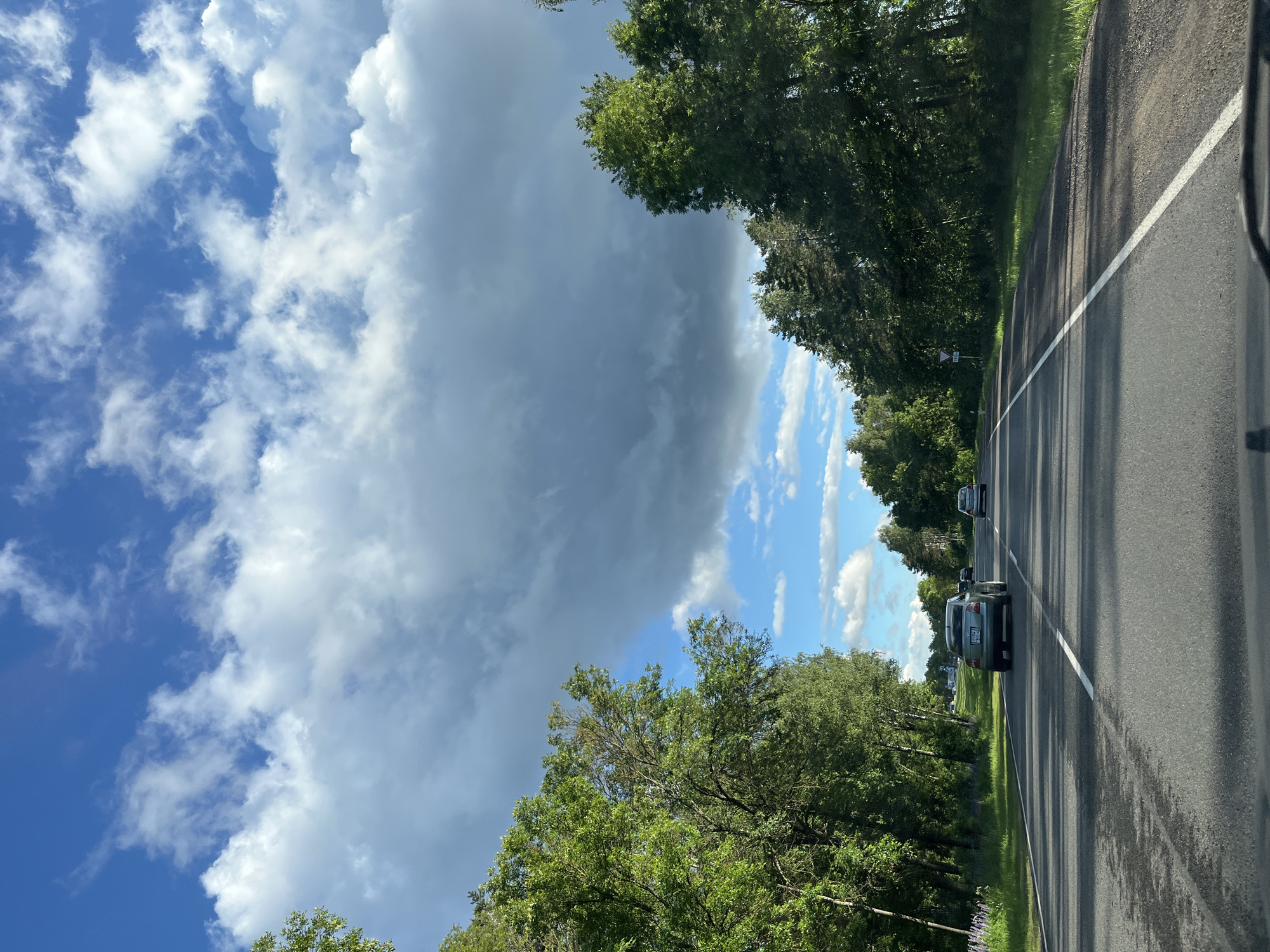 The image depicts a view from a vehicle driving on a paved road lined with trees on both sides. The road has a white line marking its edge. Several cars are visible on the road ahead. The sky is filled with a mix of dramatic, dark storm clouds and brighter, scattered white clouds against a blue sky. Sunlight filters through, casting shadows from the trees onto the road. The scene suggests a dynamic weather condition, possibly approaching or moving away from a storm. The lush green foliage of the trees indicates it's likely daytime during a warmer season. There is no visible text in the image. The setting appears to be a rural or semi-rural area in Belarus, given the location context.