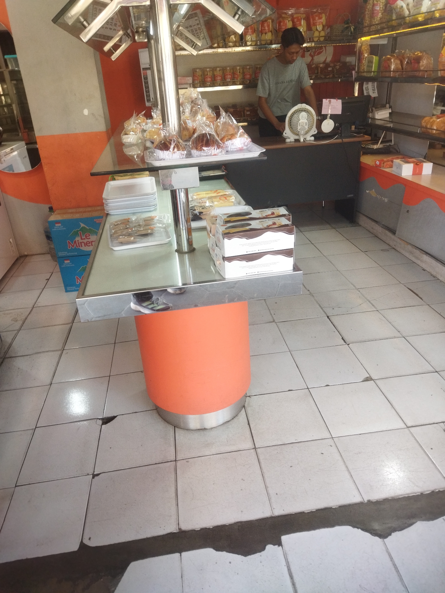 A medium shot captures a convenience store interior in Sukoharjo, Indonesia, focusing on a display table with baked goods and a counter area. A young man wearing a grey t-shirt stands behind the counter, looking down at a computer monitor. A white electric fan is positioned on the counter in front of him. The display table in the foreground is made of glass and metal, holding stacks of white plastic plates, packaged baked goods, and what appear to be small, individually wrapped snacks. Behind the counter, shelves are stocked with various food items, including jars of what seem to be preserves or snacks and bagged baked goods. The floor is tiled with white squares, some of which show signs of wear and damage. The walls are painted with an orange and grey color scheme. The overall impression is that of a small, local retail establishment.
