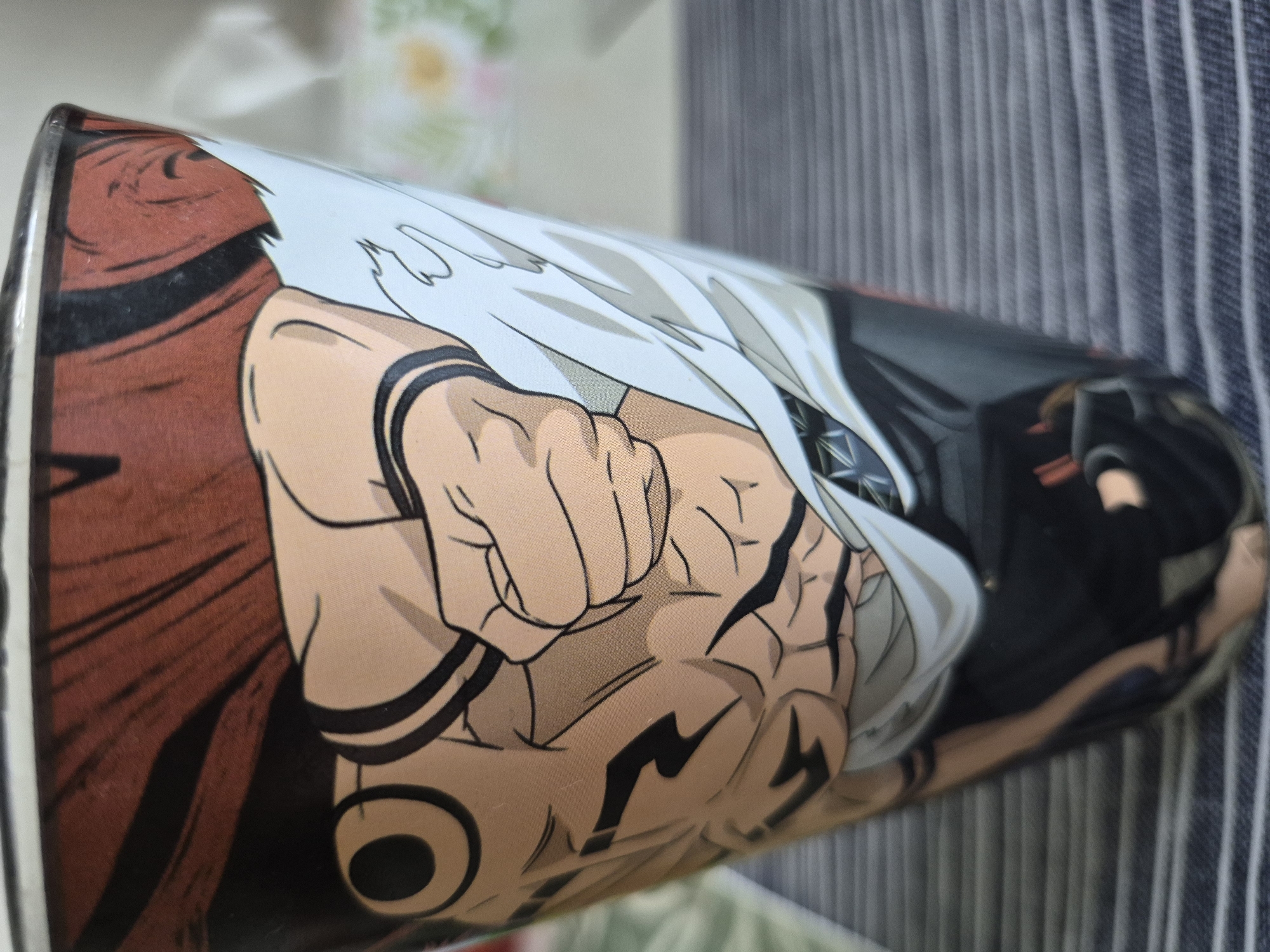 A close-up shot shows the side of a cylindrical glass. The glass is decorated with an anime-style illustration of a muscular character. The character is depicted with a clenched fist, and portions of their torso and arm are visible. The illustration features bold black outlines, flat colors, and shading to create a sense of muscle definition. The character is wearing a white garment with black stripes, and a dark red cloth is visible at the top of the glass. The background is blurred, suggesting a domestic setting, with a striped surface and hints of greenery. The scene is brightly lit, casting some shadows.