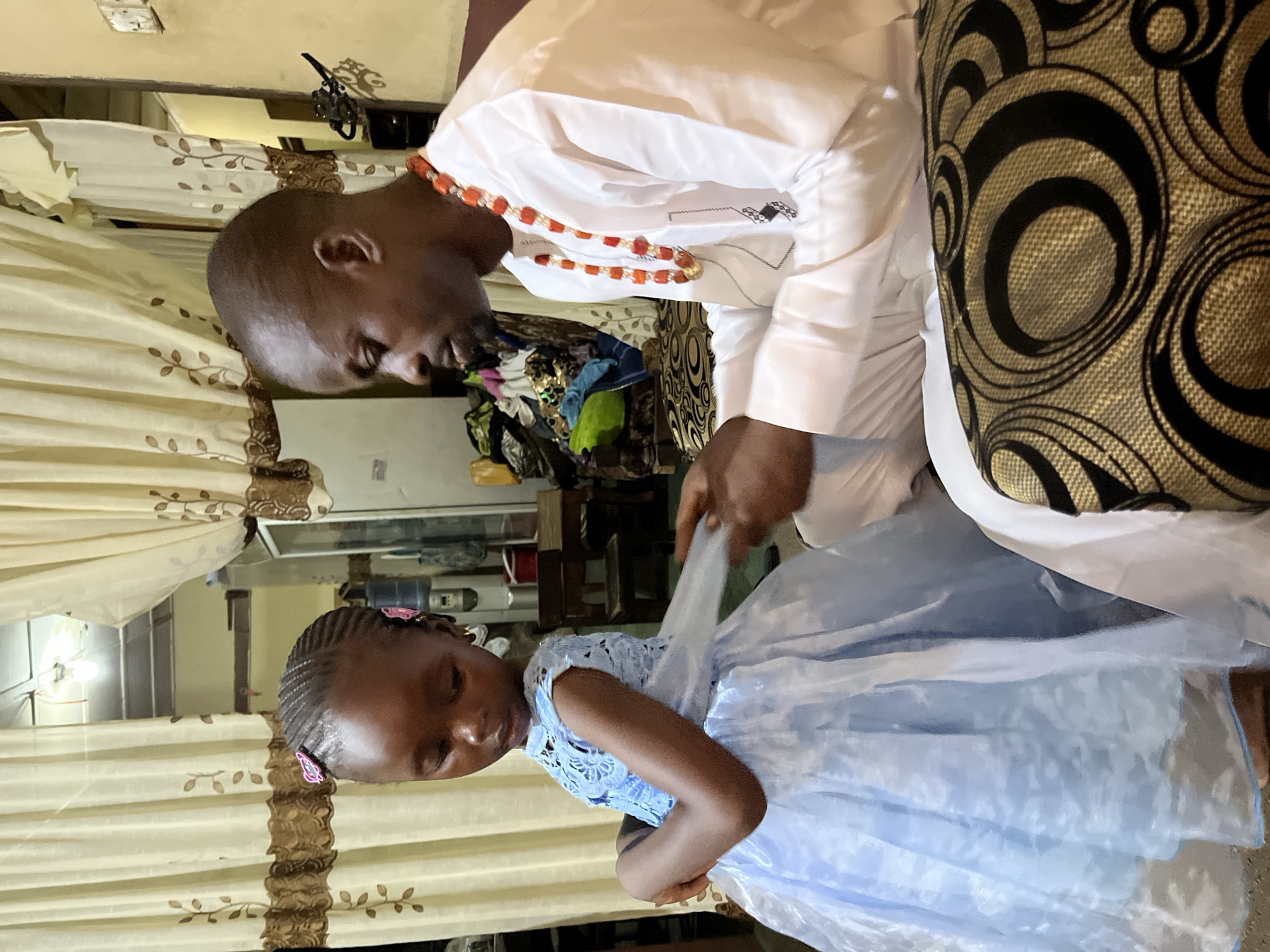 A man and a young girl are indoors. The man, facing left, is wearing a white shirt and an orange beaded necklace. He is adjusting the girl's light blue dress. The girl, also facing left, is wearing hair clips. The setting appears to be a home interior, with curtains and furniture visible in the background. There is no specific information in the image to verify the city of Rafin Bauna, Nigeria.