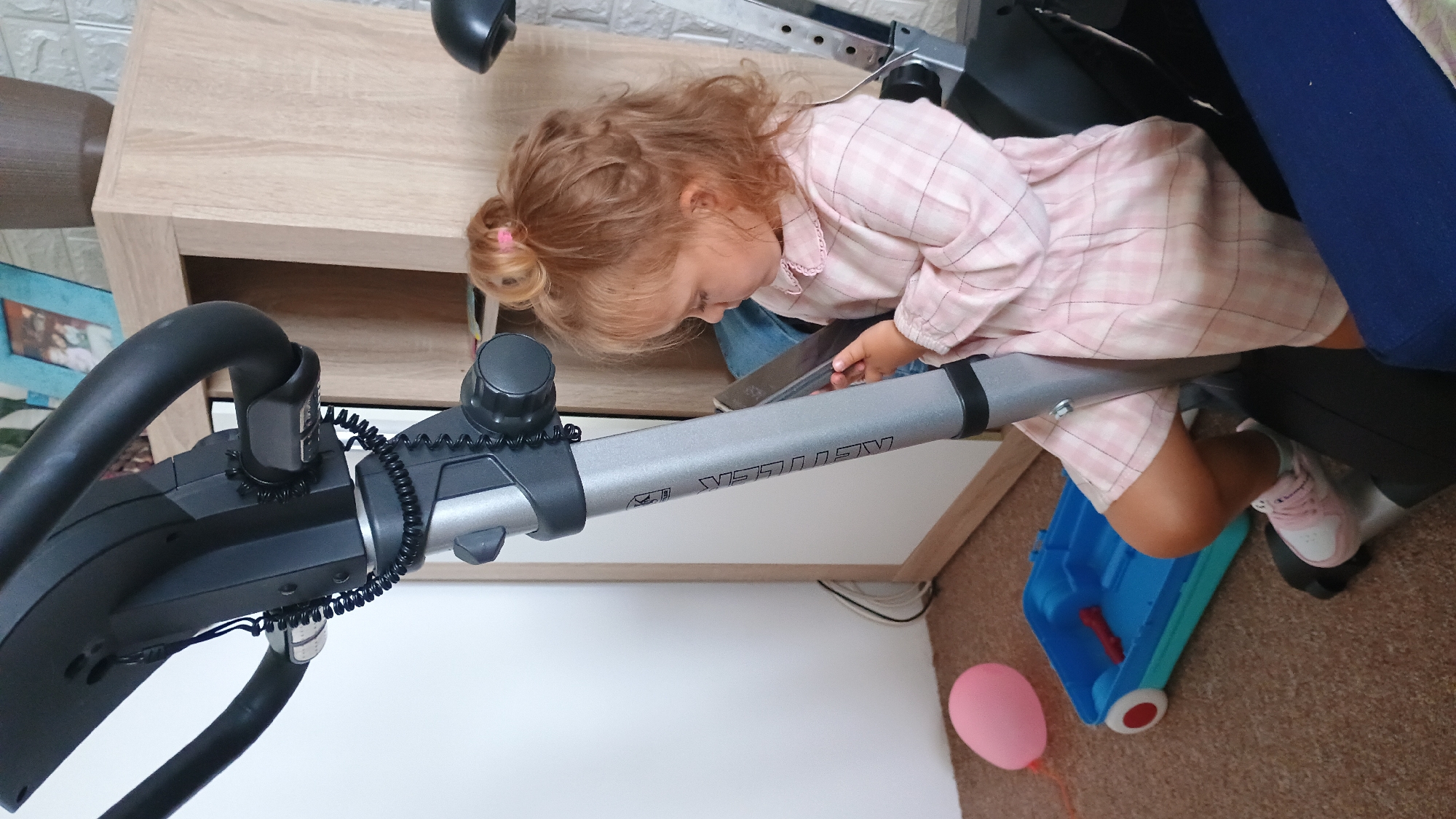A young Caucasian girl with blonde hair tied in a bun, wearing a pink checkered dress and pink and white sneakers, sits on a stationary exercise bike. She is holding a smartphone in her hands and appears to be looking at it. The exercise bike is black and gray, with a silver upright pole. To the left of the bike, on the carpeted floor, is a pink balloon and a blue plastic toy wagon with red wheels, containing a red object. In the background, a wooden cabinet with shelves is visible, and on one shelf, a framed picture and a brown planter are present. The wall behind the cabinet appears to be tiled. The scene suggests an indoor residential setting.