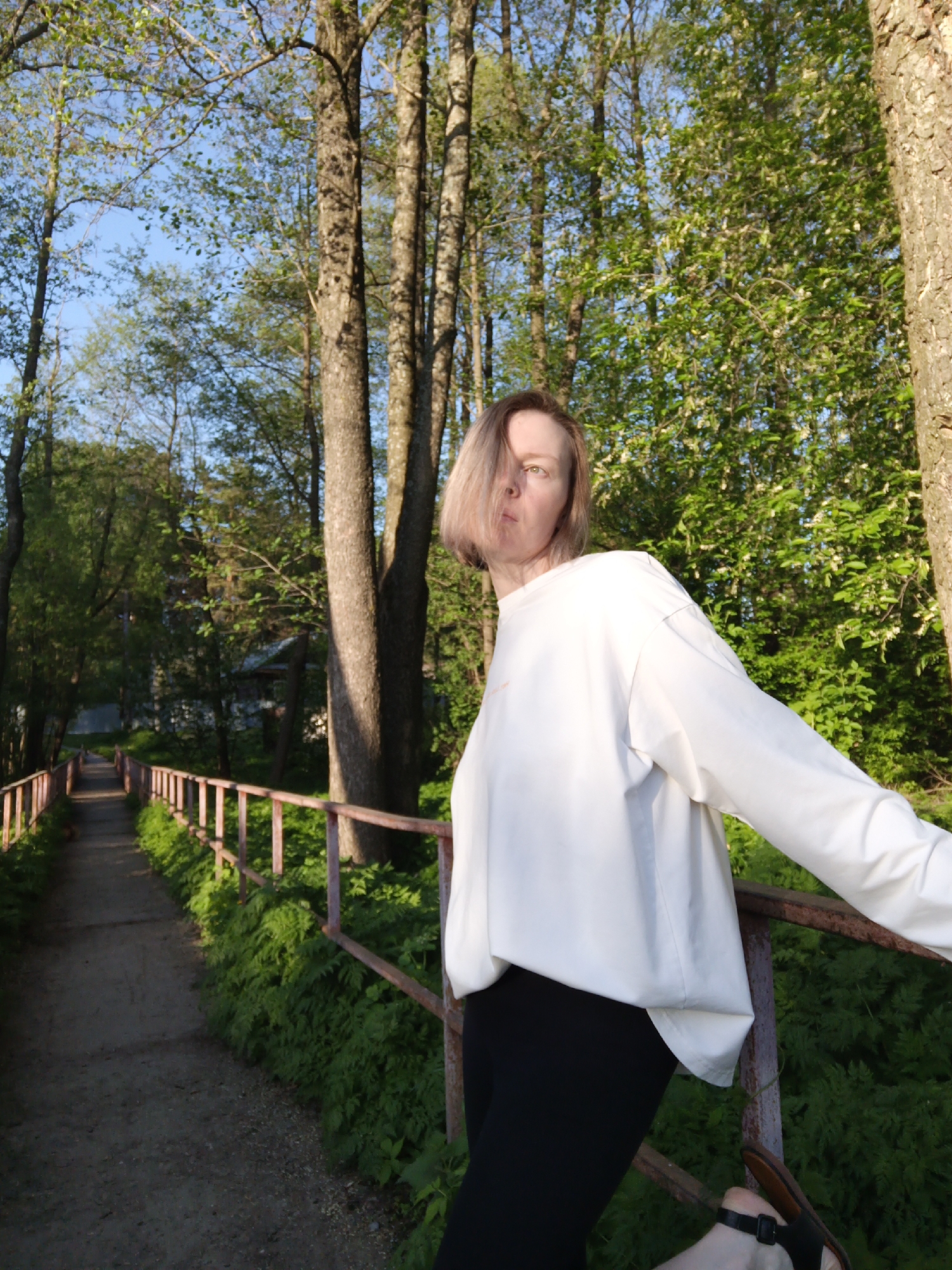 A woman stands on a bridge railing in a wooded area in Chaikovsky, Russia. She wears a white long-sleeved shirt and black pants with a sandal on her foot. The bridge has a low railing with green vegetation growing along its edges. A path stretches into the distance. Trees surround the area. The sky is visible through the trees.