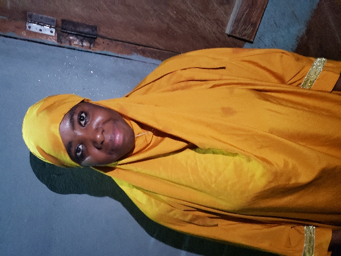 A young Black woman smiles wearing a bright yellow headscarf and gown with gold trim. The fabric drapes around her head and shoulders. She is indoors, with a plain wall behind her. Visible in the background are a brown door and some metallic hinges. The lighting in the room is soft and muted. The image is taken from a slightly high angle and rotated counterclockwise.