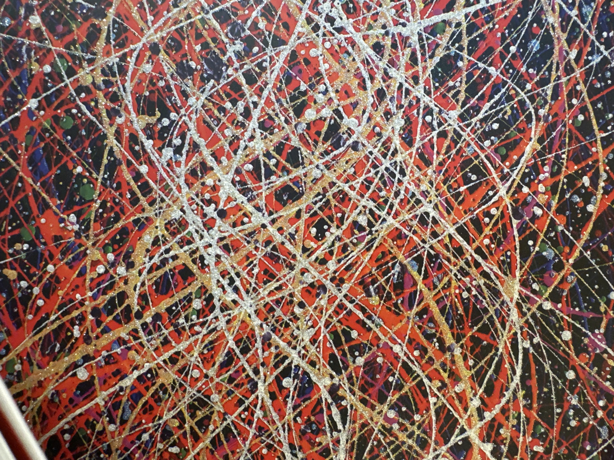 This media file is a close-up, abstract image of a painting. The painting features a chaotic splatter pattern of various colors, predominantly red, black, gold, and white. Thin lines of gold and white paint are splattered and dripped across a background that transitions between bright red and deep black. Small flecks of silver glitter are visible, catching the light. There are also hints of other colors like green, blue, and purple interspersed within the splatters. The texture of the paint appears to be layered and somewhat raised in places due to the dripping technique. There are no discernible subjects like people, objects, or a specific scene. The image captures a moment of dynamic artistic expression through paint application. No text is visible in the image, and there are no cues related to time of day, weather, or location that would suggest Minsk, Belarus. The overall impression is one of energetic and vibrant abstraction.