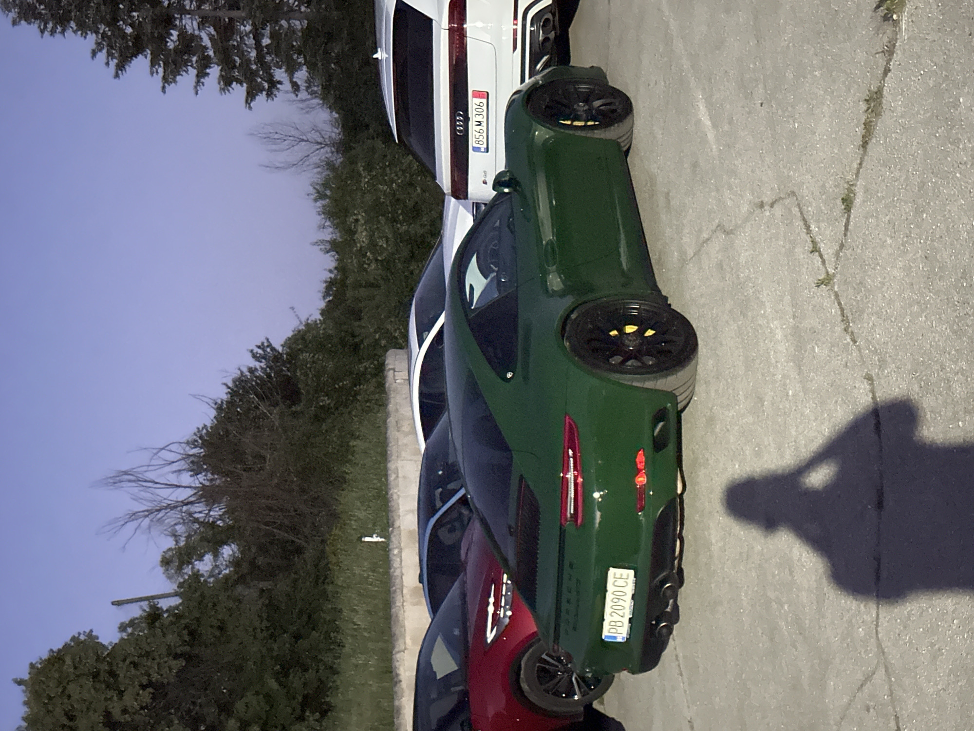 In Arbanassi, Bulgaria, a dark green Porsche 911 is parked on an asphalt surface. The car has a license plate that reads "PB 2090 CE." To the left of the Porsche, a red car is partially visible. Behind the Porsche, a white Audi SUV is parked, displaying a license plate that reads "856 M 306." The scene is set against a backdrop of trees and a clear sky, suggesting it is either early morning or late evening. A shadow of a person holding a phone is cast on the ground in front of the Porsche.