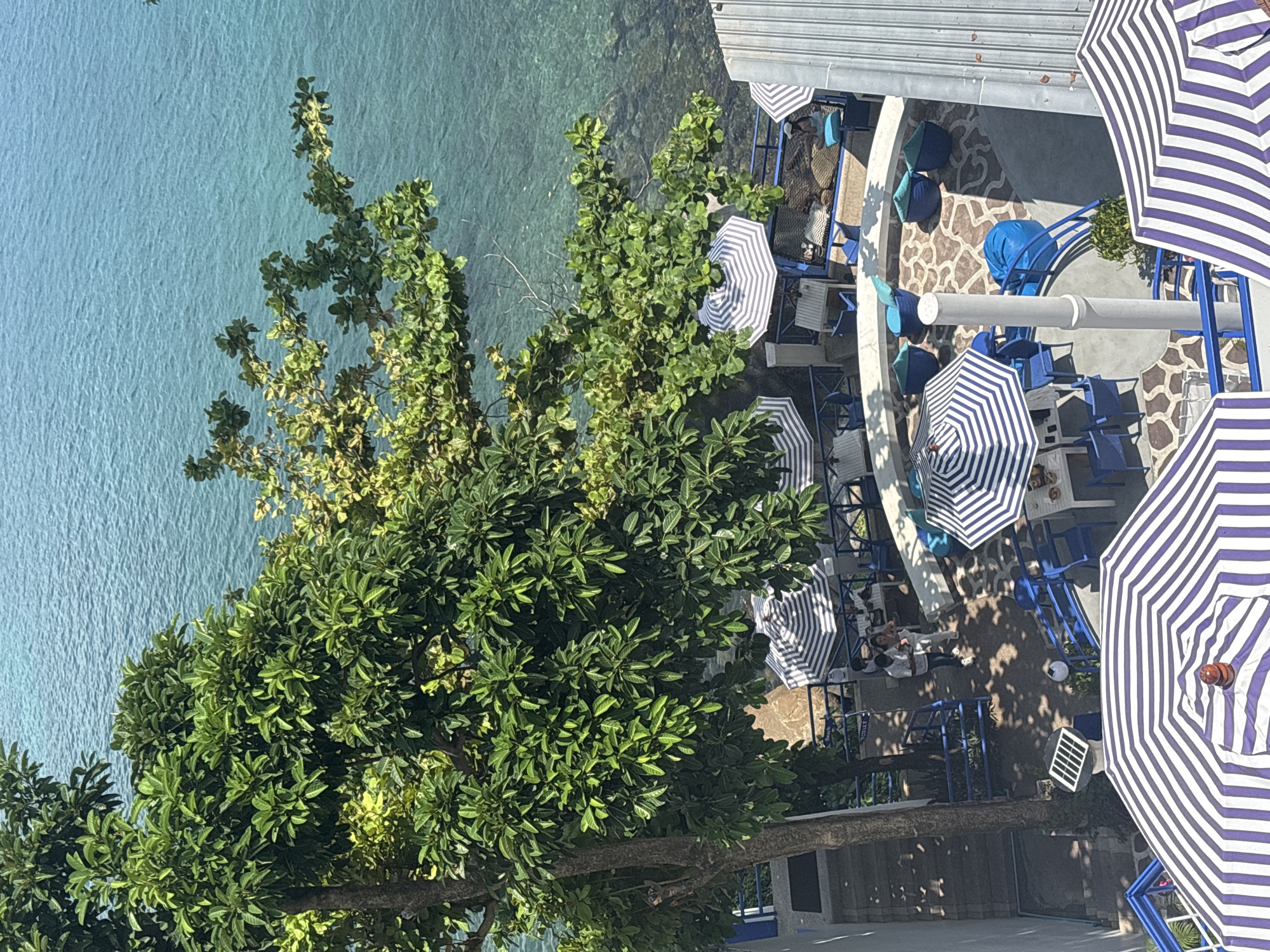 Viewed from above, the image shows a section of Kamala, Thailand. Part of the turquoise ocean is visible on the left, separated from a waterfront patio by lush green trees. The patio area, constructed with white concrete and blue railings, features blue and white striped umbrellas shading tables and chairs. Some people are seated at the tables under the umbrellas. The overall atmosphere appears relaxed and vacation-oriented.