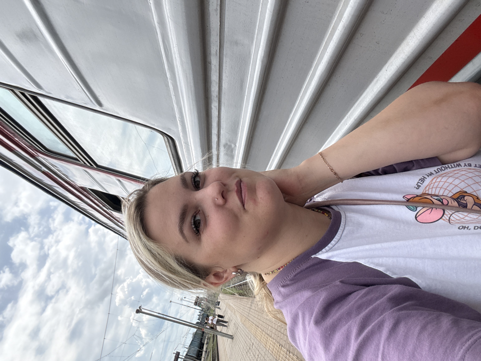 A young woman with blonde hair is taking a selfie on a train platform in Povorino, Russia. She is wearing a purple and white raglan shirt and a necklace with colorful beads. She has a nose ring and hoop earrings. Behind her, a train car with visible windows and red trim is parked. The train platform has yellow tactile paving and in the distance, other passengers can be seen walking. The sky is cloudy.