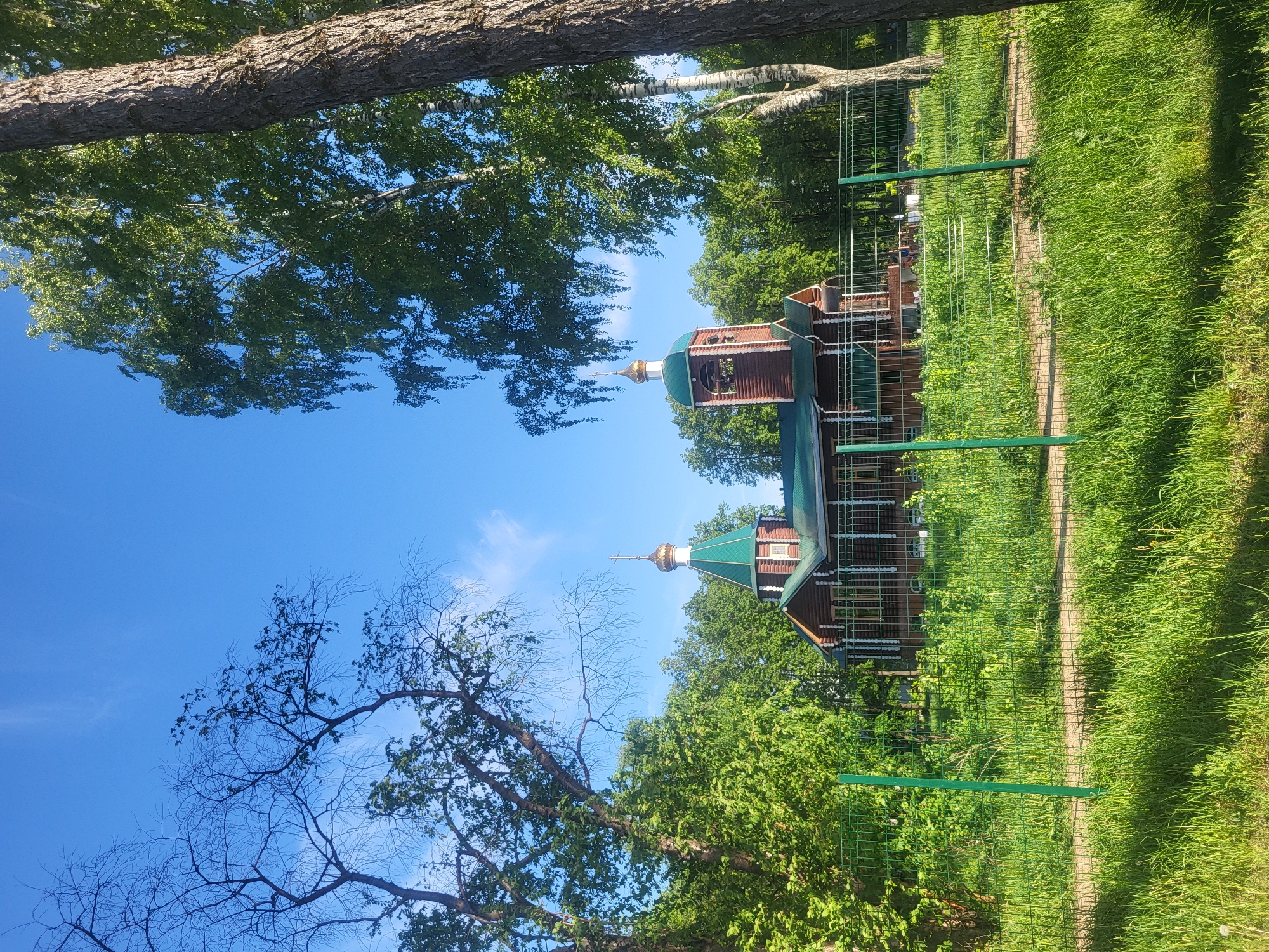 The image depicts a small wooden Orthodox church in a green, park-like setting. The church has a green roof and gold-colored domes with crosses. It's surrounded by trees and tall green grass. A green metal fence runs along the perimeter. The sky is blue with scattered clouds. The picture is taken on a sunny day. The city is Chaikovsky, Russia.
