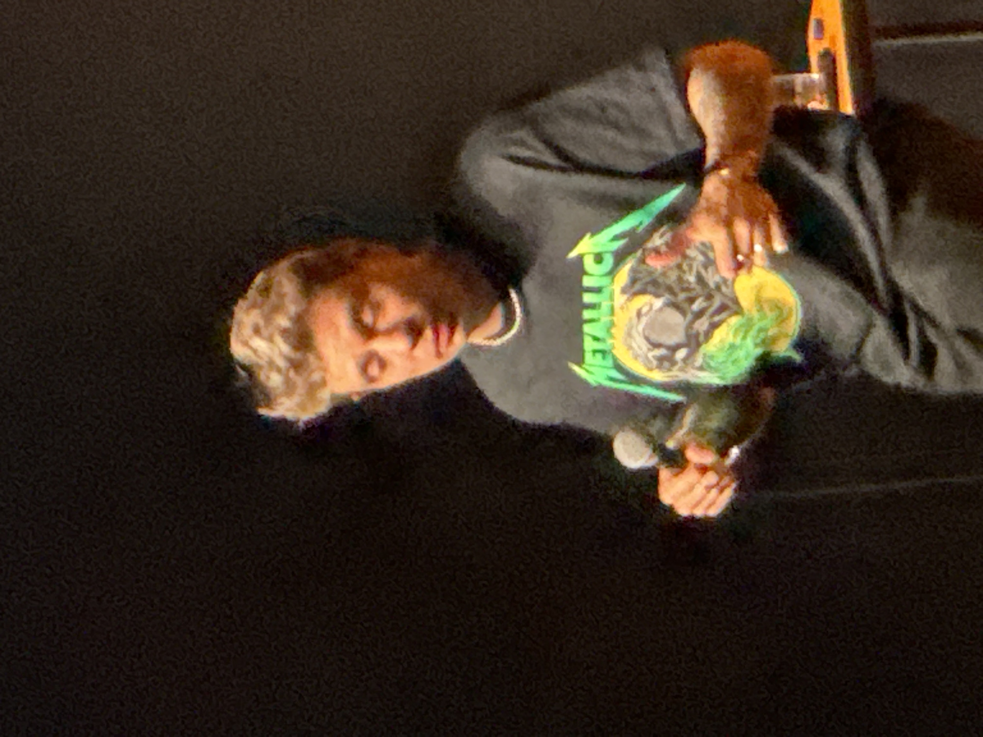 The image displays a young man performing on what appears to be a dimly lit stage. He has light brown, wavy hair and is wearing a dark grey, long-sleeved t-shirt. A prominent graphic on his shirt features the word "METALLICA" in neon green text, situated above a detailed illustration of a skull or monster-like figure in yellow, green, and grey tones. He is also adorned with a white or silver chain necklace.
The man holds a microphone in his right hand, with his left hand also visible, possibly supporting or gesturing. His eyes are partially closed or looking downwards, and his mouth is slightly open, indicating he is actively singing or speaking. The background is uniformly dark, suggesting a concert or performance setting, with a blurry hint of a musical instrument or stand visible in the upper right corner. The overall scene conveys a focused performance in an indoor venue, located in Vnukovo, Russia.