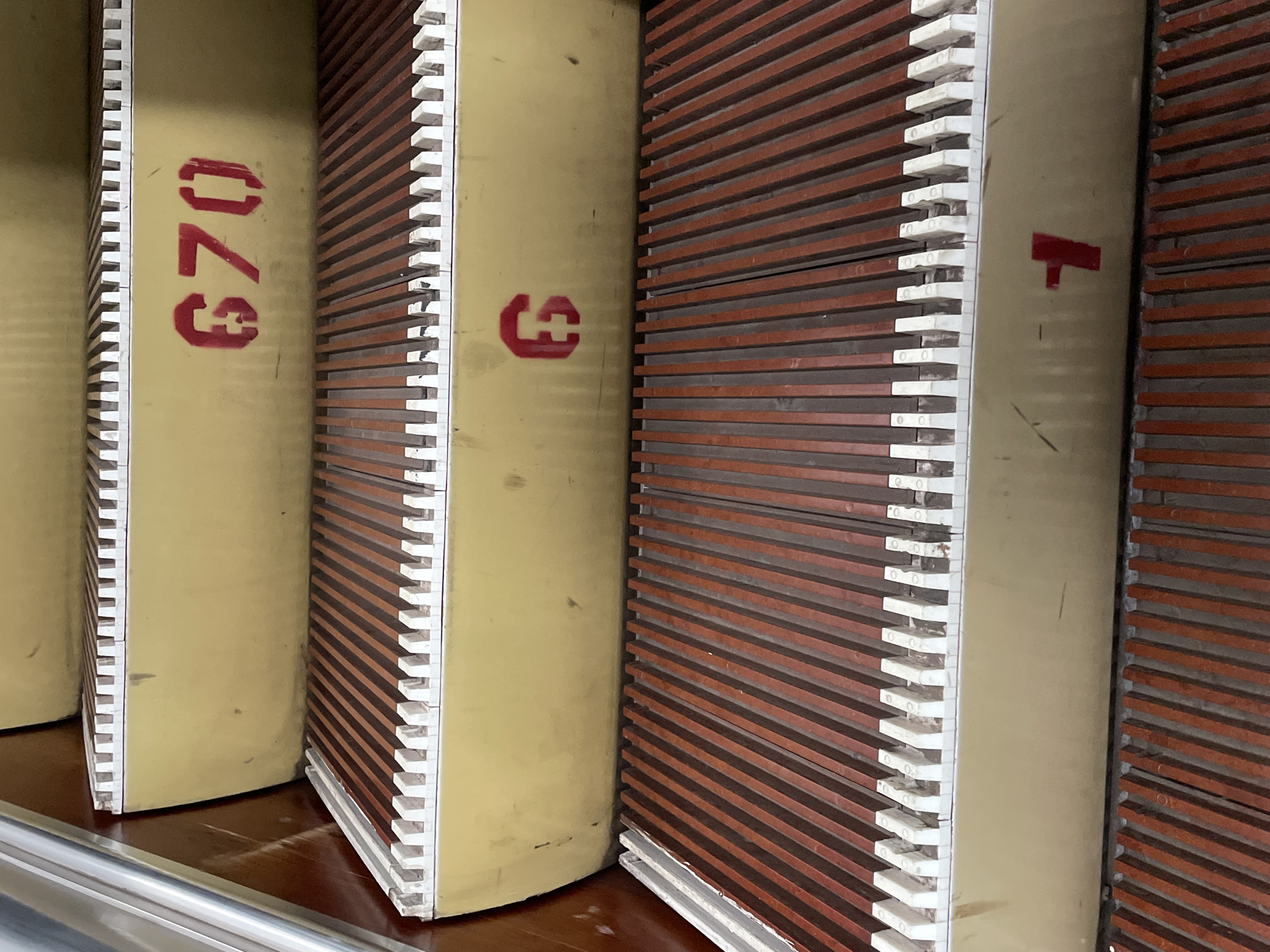 An upward view of an escalator shows four steps. The steps are made of alternating brown and dark brown ribbed metal. The front edge of each step has a white comb-like tread. The risers of the escalator are a muted yellow color. Stenciled in red on the riser of the second step from the top is the number "670." On the riser of the third step from the top is the number "6." On the riser of the fourth step from the top is the number "1." The left side of the escalator has a brushed metal handrail.