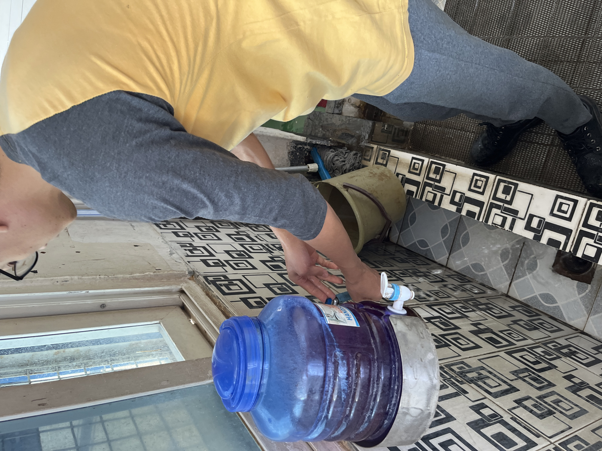 A person in a yellow and grey raglan t-shirt and grey sweatpants is filling a blue bucket with water from a large blue water jug. The water jug is on a metal stand attached to a tiled wall. The tiles have a black and white geometric pattern. The person's hands are near a white faucet attached to the water jug. There is a dirty mop and a second bucket near the person. The scene appears to be in an outdoor or semi-outdoor area with a window and a drain in the floor. The location is identified as Pelang, Indonesia.