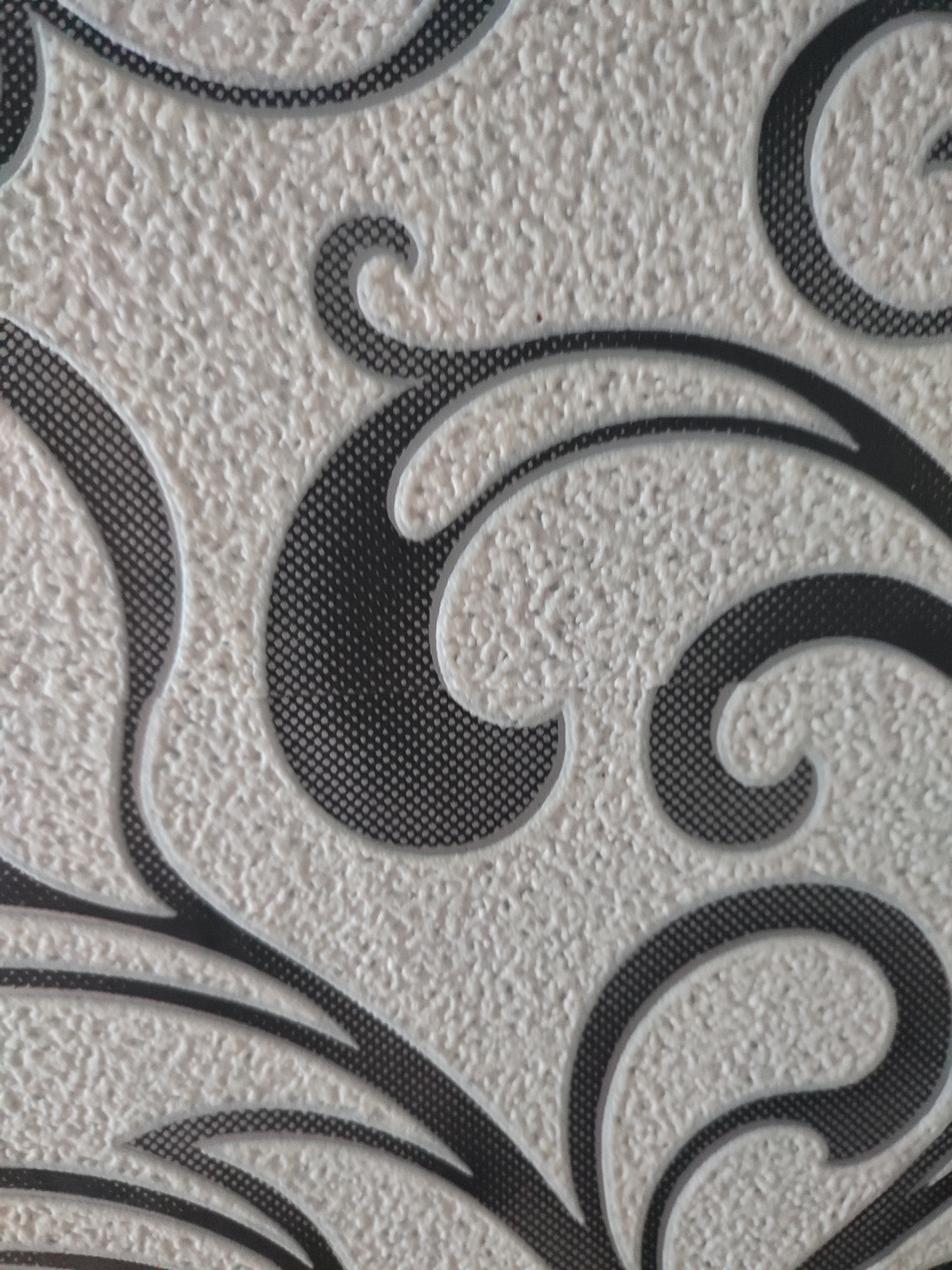 The image shows a close-up of a textured, light-colored surface with a repeating black and grey scroll pattern. The texture appears to be a raised, stippled effect. The pattern consists of swirling lines and curved shapes, some filled with a grey dotted pattern, and outlined in black. The lighting is even, with no harsh shadows, suggesting an indoor or overcast environment. There are no people, animals, or distinct objects visible beyond the patterned surface itself. The location in Narowlya, Belarus, is not discernible from the visual information in the image.