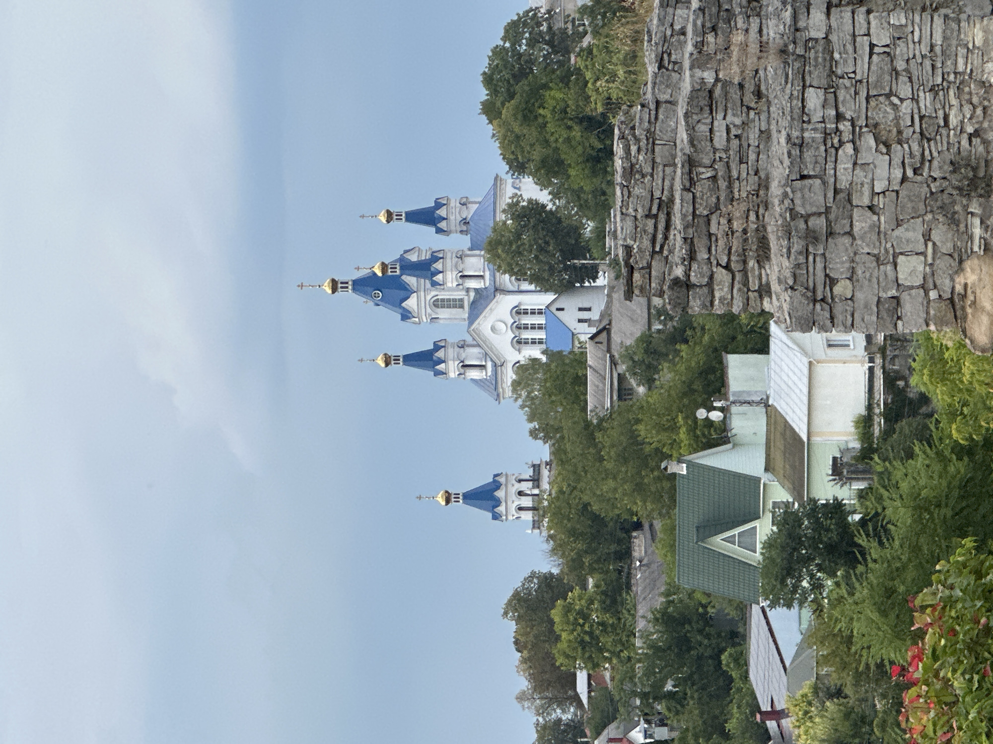 The image depicts a panoramic view of Kamianets-Podilskyi, Ukraine, featuring a prominent Orthodox church with blue domes and gold spires, surrounded by greenery and residential buildings. In the foreground, a stone wall, likely part of the ancient Kamianets-Podilskyi Castle, occupies the lower right portion of the frame. The sky is overcast with light blue clouds. The scene captures a blend of historical architecture and everyday life within the city, with the church serving as a focal point against a backdrop of trees and rooftops.