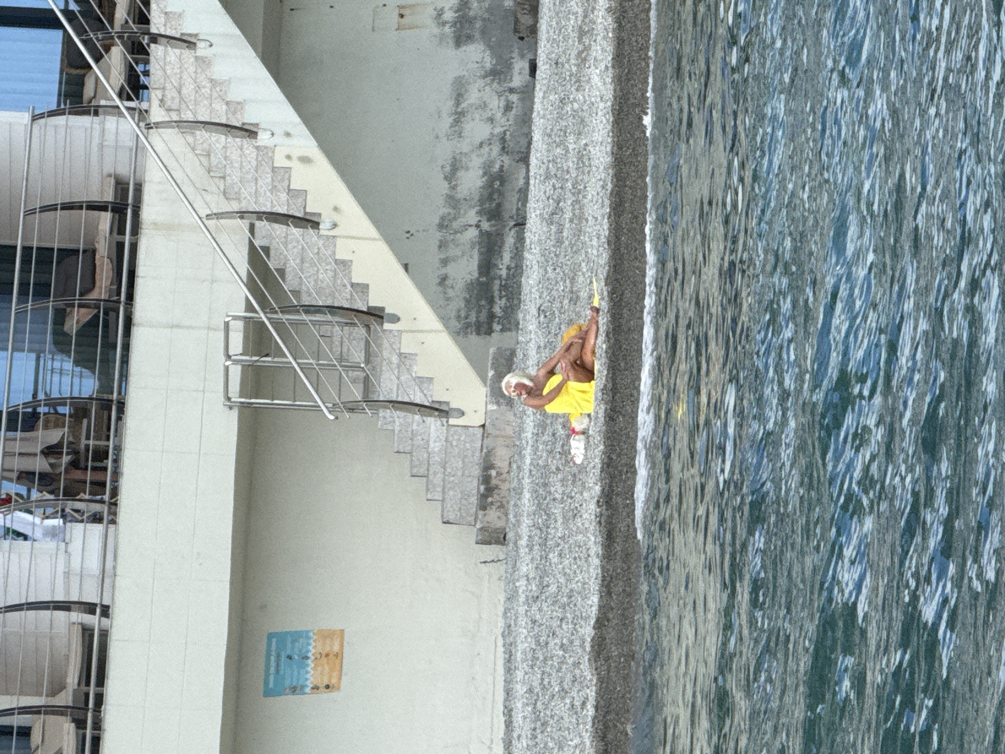 A person in a yellow top and white shorts is sitting on a pebble beach next to a body of water. The person has blonde hair and is wearing white shoes. The water appears to be a light blue color with ripples on the surface. To the left of the beach is a large, light-colored building with a set of stairs leading down to the beach. The stairs are made of stone and have metal railings. The building has several windows and a modern design. A small, colorful artwork is visible on the wall of the building. The scene appears to be outdoors during the daytime, with natural light illuminating the area. The overall impression is one of a relaxed and leisurely setting, possibly a beach resort or a public recreational area. The orientation of the image is sideways.