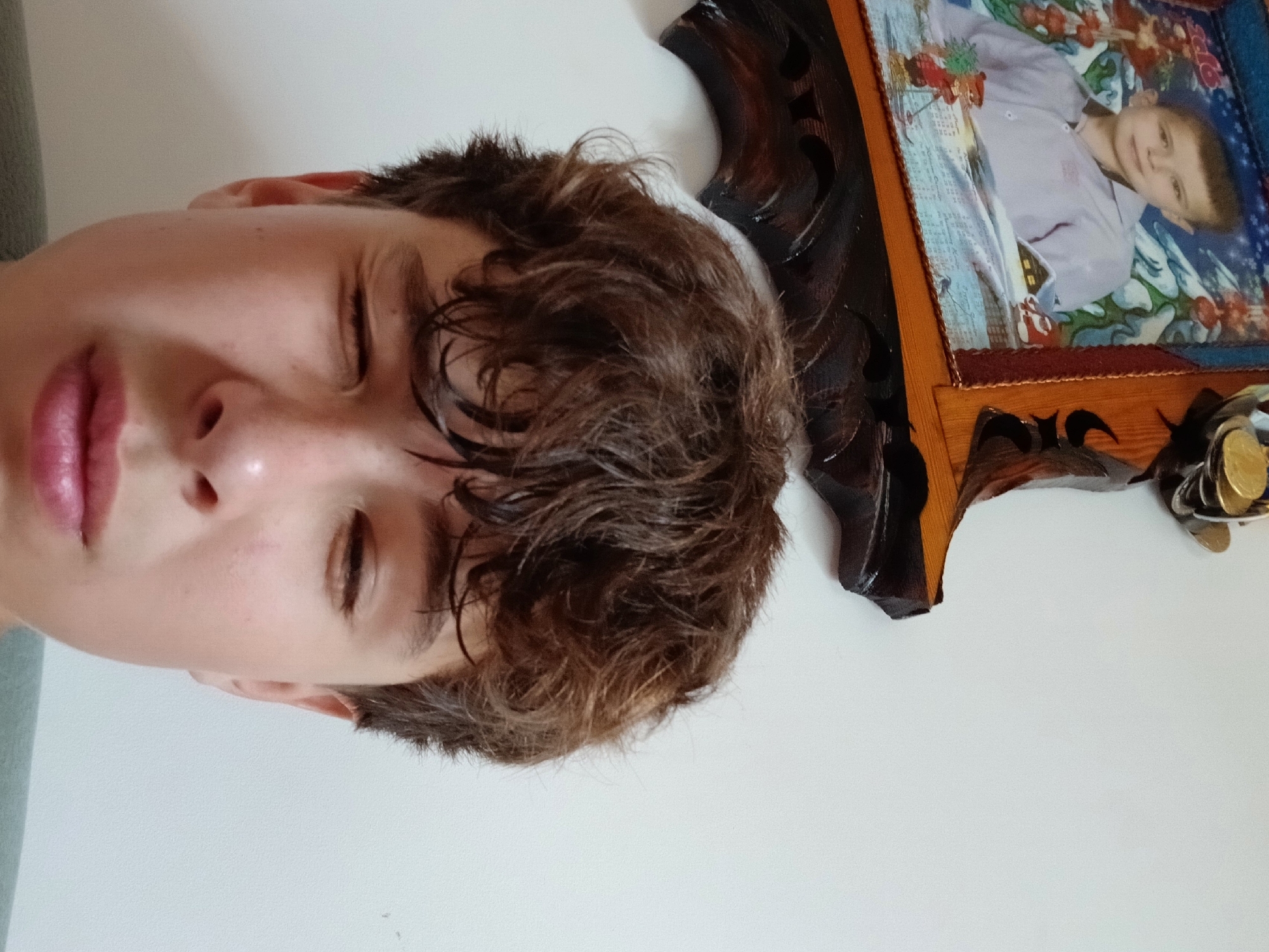 A close-up selfie of a young male with brown, curly hair is captured against a plain white wall. The individual is looking downwards and to the left, with his left eye closed and his right eye squinting. His mouth is slightly open, revealing his teeth. To the upper left of the frame, a framed photograph is visible. The photograph appears to be of a young boy dressed in a light-colored collared shirt, set against a wintery backdrop with snow-covered mountains and a small cabin. The frame itself is ornate and dark brown, with carved details. The bottom portion of the frame is out of focus. The scene is indoors, with the white wall suggesting a domestic interior. There are no discernible elements within the image that indicate a specific location in Dnipro, Ukraine.