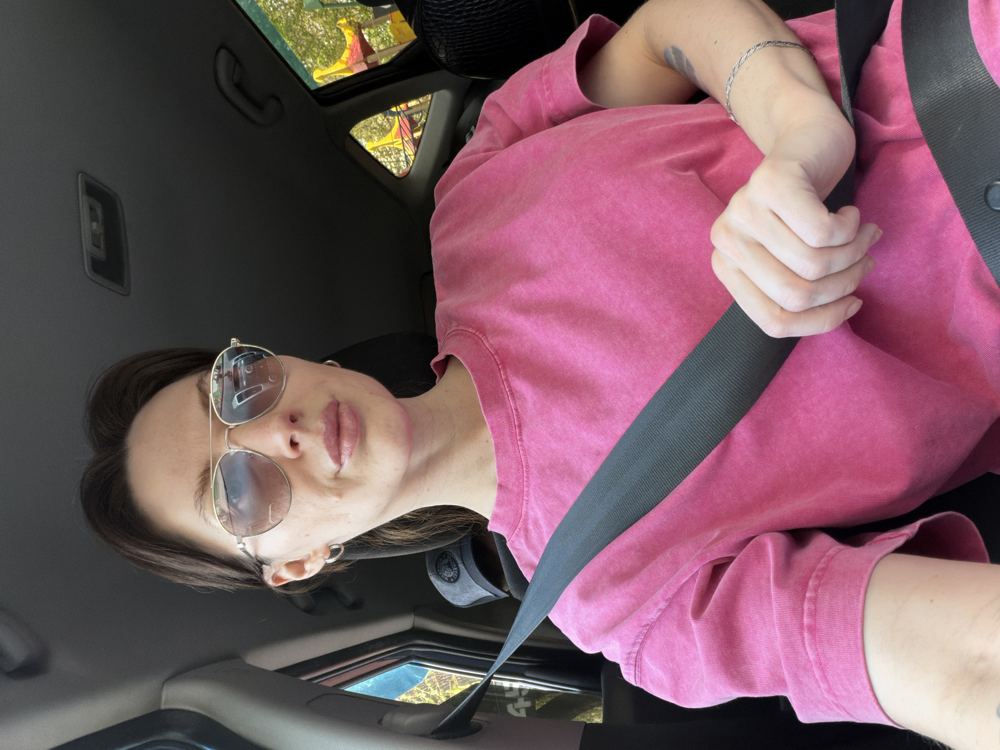 A person is shown from the chest up, sitting in the driver's seat of a car with a seatbelt fastened. The person is wearing a pink t-shirt and round-framed sunglasses, and has short brown hair. A silver bracelet is visible on their left wrist. The car interior is gray and black. Through the car's sunroof, a view of trees and a yellow and red playground structure is visible. The location is difficult to determine with certainty based on the given information. Perm, Russia, cannot be verified as the location from the image alone.