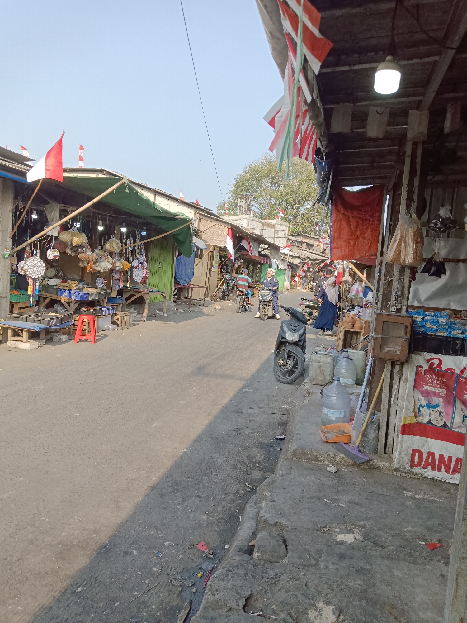 The image depicts a street scene in Waringinjaya, Indonesia, likely a market area. The street is paved and bordered by informal stalls and buildings constructed with corrugated metal and wood. Red and white Indonesian flags are prominently displayed on several structures, suggesting a national holiday or celebration.
In the foreground, to the right, a motorbike is parked on the side of the road. In front of a stall, several large water jugs and cleaning supplies, including a broom and a dustpan, are visible. A wooden cabinet is mounted on a pole. A large banner with the word "DANA" in red letters is also displayed.
Further down the street, several people are present. A woman in a blue headscarf and dress is walking towards a stall. Another person, possibly a man, is standing near her. Two individuals on motorbikes are also visible further down the street, with one person riding and another appearing to be a passenger.
The stalls along the street are filled with various goods, including what appear to be packaged food items, decorative items, and possibly household goods. Some stalls have green awnings or tarps providing shade. The overall environment suggests a bustling commercial area with a rustic and informal setup. The sky is clear and a pale blue.