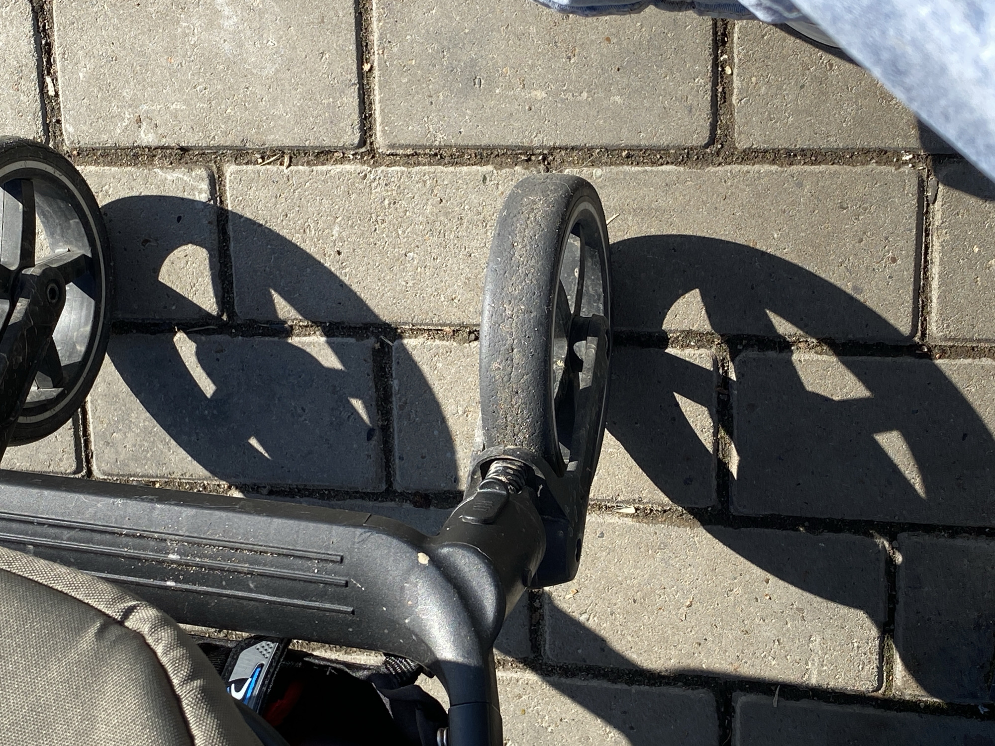 The scene, located in Chaikovsky, Russia, depicts an overhead view of an outdoor paved area under direct sunlight. The primary subjects are components of a wheeled device, likely a stroller or similar transport mechanism. A segment of grey canvas fabric is visible on the upper left, attached to a vertical, ribbed black plastic structural element. This element connects to a black plastic housing, which supports a swiveling wheel positioned centrally in the frame. This wheel features a worn, dark grey tire and black spokes. A second wheel, also with black spokes and tire, is partially visible at the top of the image. The ground is composed of light grey rectangular concrete pavers, with visible dirt, small debris, and sparse organic matter within the grout lines. Strong sunlight casts distinct, dark shadows of the wheeled device's components and wheels onto the pavement. On the far right, a portion of a human leg, clad in light blue jeans, and dark footwear are visible, indicating the presence of an individual standing adjacent to the wheeled device.