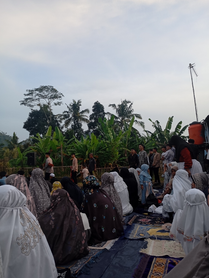 The image shows a group of people gathered outdoors in a clearing surrounded by trees and foliage. They are all wearing traditional Islamic clothing and some are kneeling, praying, or sitting on the ground. There are a few prayer rugs spread out, and the people are facing towards the left of the frame. The scene is set in an open-air area with a lush green background, possibly a mosque courtyard or a garden. There is a blue plastic tarp covering the ground. The overcast sky suggests it is early morning or late afternoon. The location based on the provided coordinates is in the village of Kutawaringin, Indonesia.