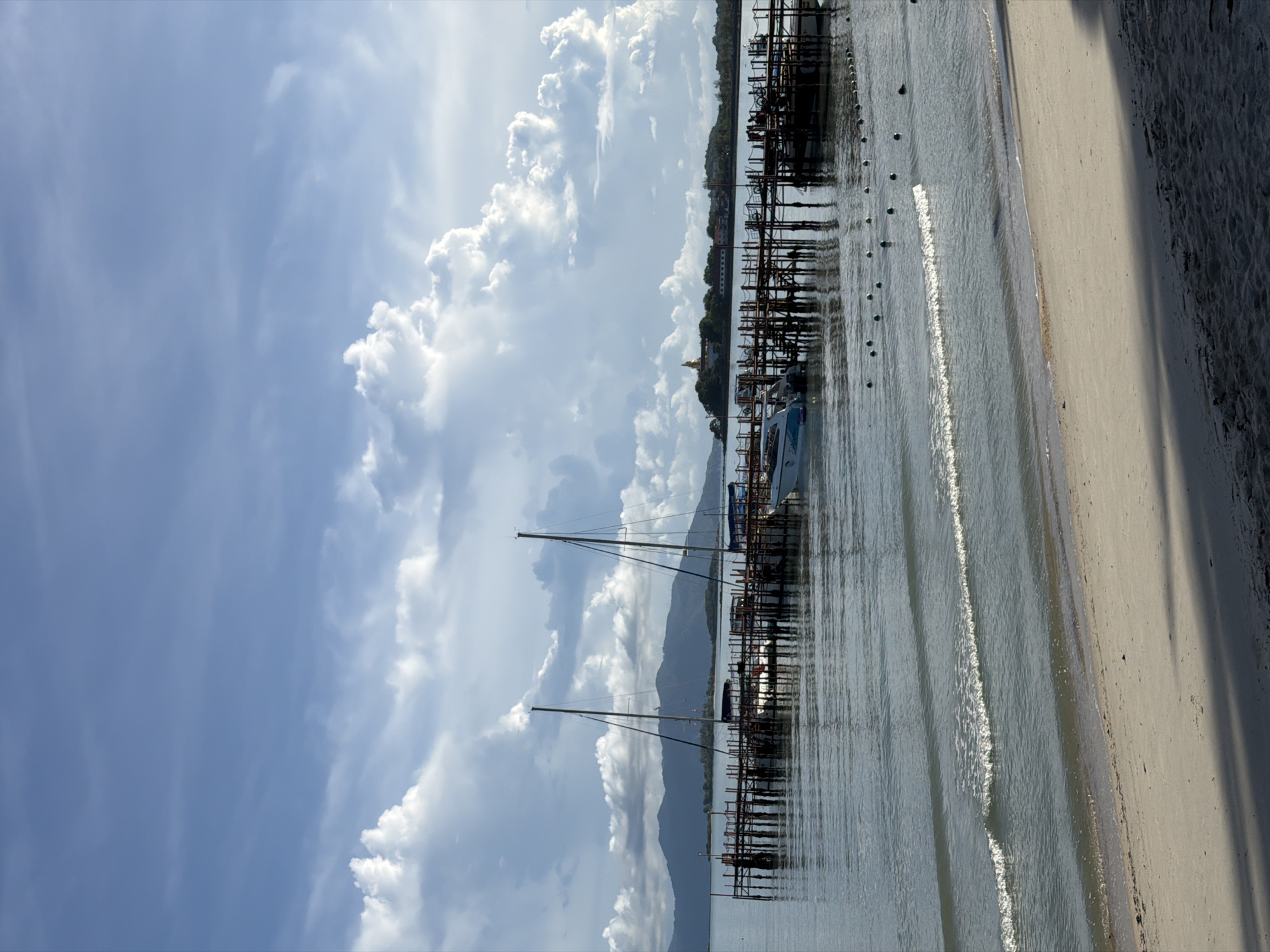 The image depicts a scene at the Baan Bang Rak pier in Thailand. The foreground shows a sandy beach with gentle waves lapping the shore, casting shadows from unseen objects onto the sand. In the mid-ground, a wooden pier extends into the calm, blue water. Several boats are moored at the pier, including two sailboats with tall masts and a white motorboat. Beyond the pier, a chain of buoys floats on the water's surface. In the background, lush green hills rise against a sky filled with dramatic, white cumulus clouds. The overall atmosphere is serene and picturesque, characteristic of a tropical coastal setting.
