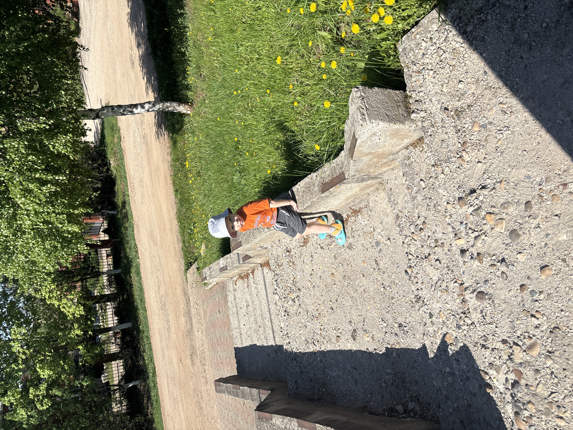 A young child lies on a concrete surface in Ферма, Russia. The child wears an orange shirt, gray shorts, a light-colored hat, and blue sandals. The concrete surface has a gravelly texture and is casting a large shadow. To the left is a dirt road bordered by trees and a fence. To the right is grass with yellow flowers. The image appears to be taken from an elevated position.