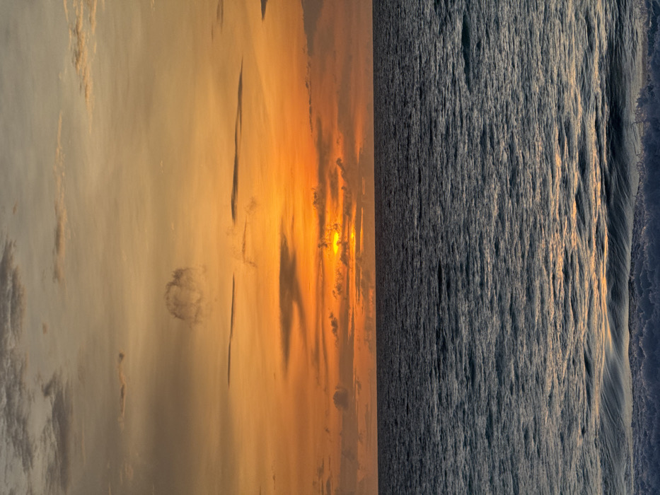 The image depicts a sunset over the ocean, presumably off the coast of Pecatu, Indonesia. The upper half of the frame is filled with a sky transitioning from a soft grey-blue at the top to vibrant orange and yellow hues near the horizon, where the sun is visible as a bright, glowing disc partially obscured by a horizontal band of clouds. Scattered, elongated dark grey clouds are present across the sky, with a distinct, somewhat circular cloud formation visible in the upper left quadrant. The lower half of the frame shows the dark blue-grey surface of the ocean, exhibiting subtle wave patterns and ripples that reflect the sky's colors. The horizon line is sharply defined and horizontal. At the very bottom of the image, the white foam and crests of breaking waves are visible in the immediate foreground. There are no people, man-made objects, or specific actions/interactions depicted in the scene.