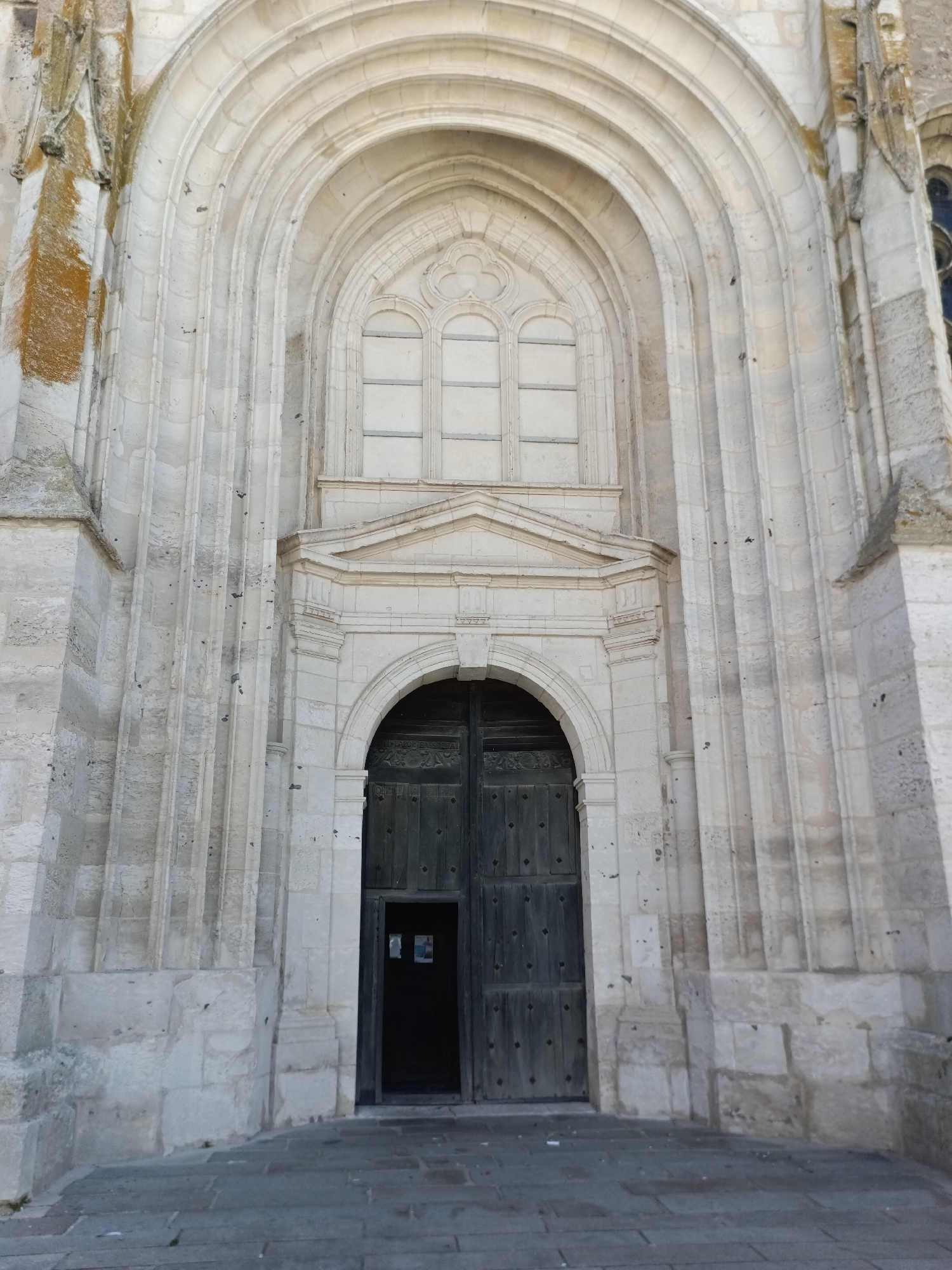 A low-angle, full outdoor shot shows the imposing stone facade of the Église Saint-Laurent in Le Neubourg, France. The central feature is a large, dark wooden double door, partially ajar, revealing a sliver of darkness within. Above the doorway, a series of concentric stone arches, characteristic of Romanesque architecture, frame a tall, narrow window with a tracery design. The weathered stone of the building displays a textured surface with some discoloration and small dark marks. The ground in front of the entrance is paved with gray flagstones. The lighting suggests it is daytime, casting subtle shadows on the architectural details.