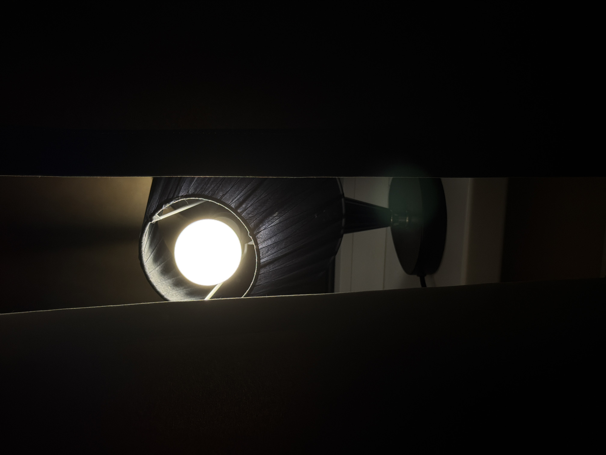 A lamp with a black shade is illuminated in a dark room, as viewed through a narrow horizontal opening. The light source is a bright white bulb. The lamp appears to be mounted on a wall, with the mounting hardware partially visible. The environment is predominantly dark, with the lamp being the primary source of illumination. It is difficult to determine the location as being in Rossiyskiy, Russia based on the provided image.