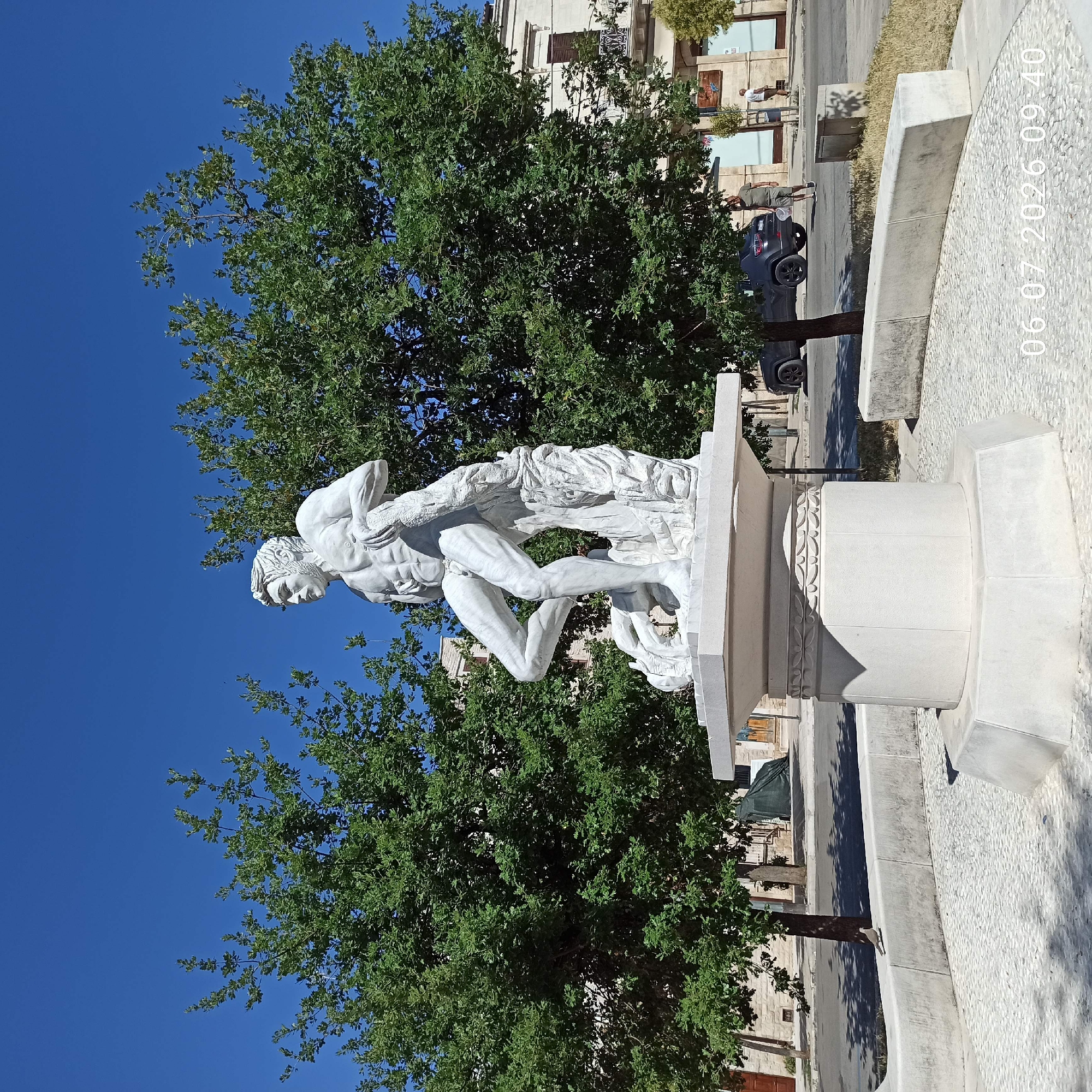 The image shows a white marble statue of a muscular man in a dynamic pose, possibly wrestling with a creature or draped in fabric. The statue is mounted on a multi-tiered, octagonal pedestal, which sits on a circular base paved with small, light-colored stones. Two large, leafy green trees flank the statue. In the background, there is a street with a parked dark-colored SUV and several people walking. A building with classical architectural elements, including arched windows and a balcony, is visible behind the trees. The sky is clear and blue. The scene is likely a public square or park in the city of Ruvo di Puglia, Italy, given the architectural style and vegetation. A timestamp "06.07.2026 09:40" is superimposed on the lower right portion of the image.