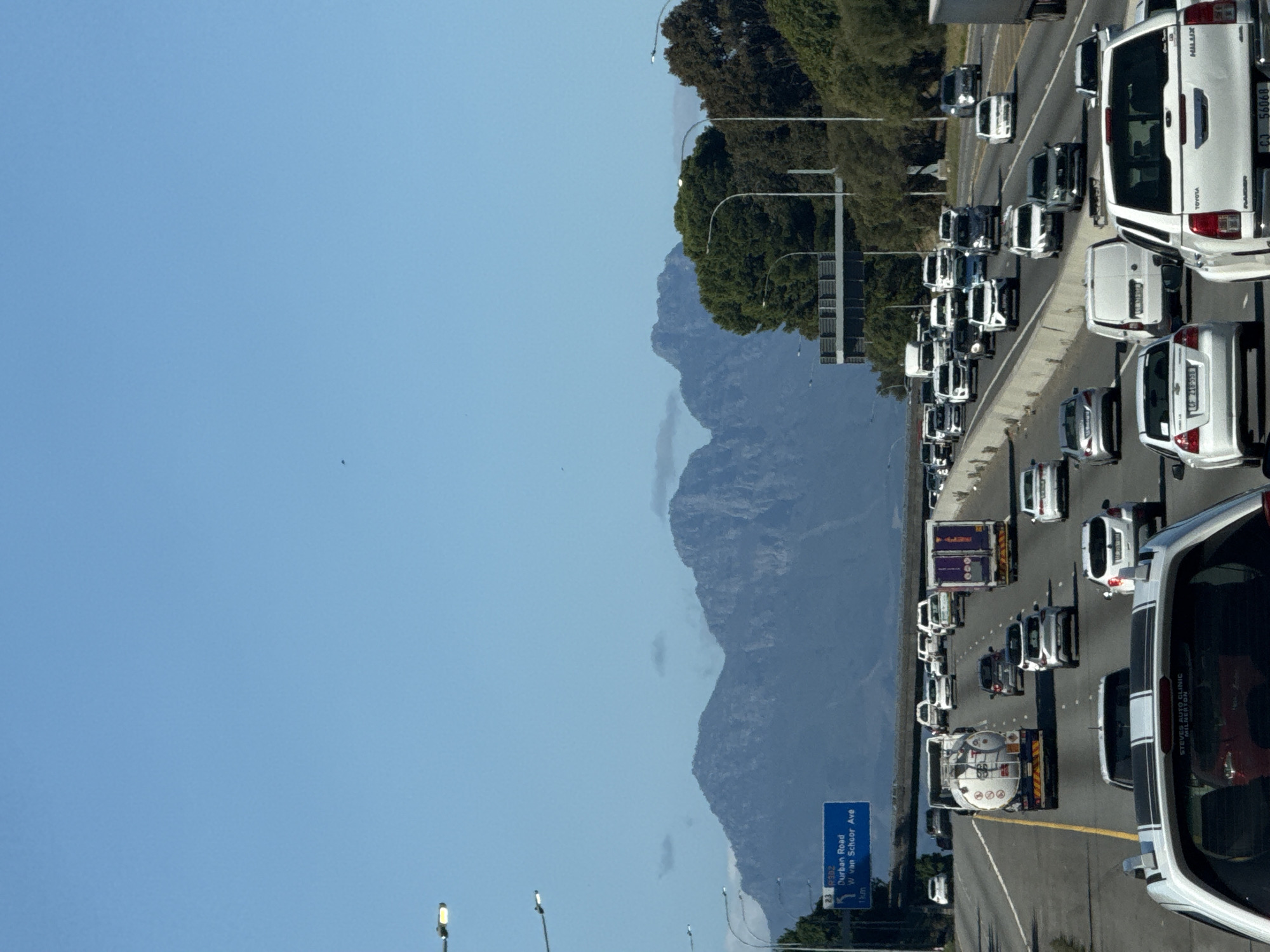 This image captures a busy highway scene, likely near Bellville, South Africa, as indicated by the visible road names and vehicle registrations. The photograph appears to be taken from an elevated perspective, looking down a multi-lane road experiencing heavy traffic.
Numerous vehicles, including cars, vans, and trucks, are visible across all lanes, suggesting either a traffic jam or slow-moving congestion. In the foreground on the right, a prominent white Toyota Hilux bakkie is seen, bearing the text "TOYOTA," "HILUX," and the license plate "CJ 56068." A smaller sticker on its rear reads "STEVES AUTO CLINIC MILNERTON." Further down the right lane, another car has the license plate "CF 218-558." On the left lanes, a tanker truck with "000" on its side is part of a queue of vehicles.
Overhead streetlights line the highway, and a large blue road sign is visible on the left. The sign provides information for upcoming exits, stating "23 R302," "Durban Road," "W van Schoor Ave," and "1km."
The background reveals a majestic range of rugged mountains under a clear, bright blue sky, typical of the Western Cape landscape. Lush green trees grow along the side of the highway, bordering the distant mountains. The scene is illuminated by strong daylight, indicating sunny and clear weather conditions. The overall impression is one of a bustling urban or suburban transport corridor during peak hours.