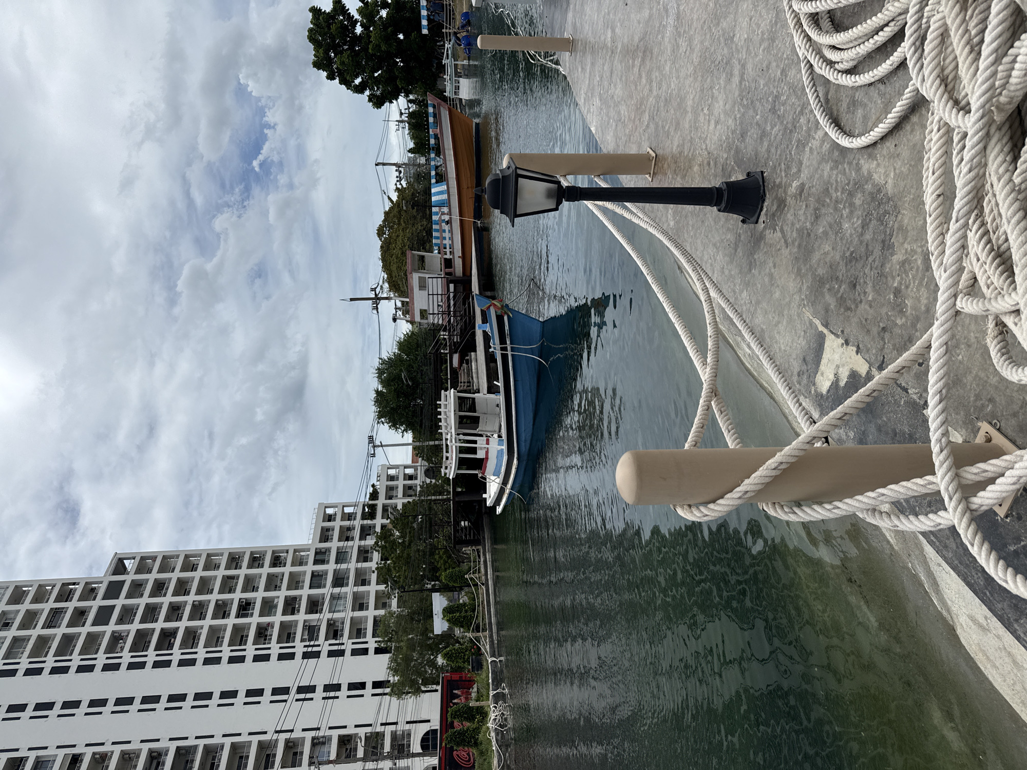 This image captures a scene at a canal in Pattaya City, Thailand, with a large, white multi-story building dominating the left side of the frame. The building features numerous balconies with dark-framed windows. In the canal, two boats are visible: a smaller blue and white boat in the foreground, and a larger orange and white boat with a striped awning further back. The canal water is dark and reflects the overcast sky.
The foreground of the image shows a concrete walkway with thick, white ropes coiled on the ground and running along a light-colored bollard, creating a nautical aesthetic. A black lamppost stands near the bollard. The background includes trees, utility poles with wires, and other structures, suggesting an urban environment adjacent to the waterway. The sky is heavily clouded with patches of blue sky visible.