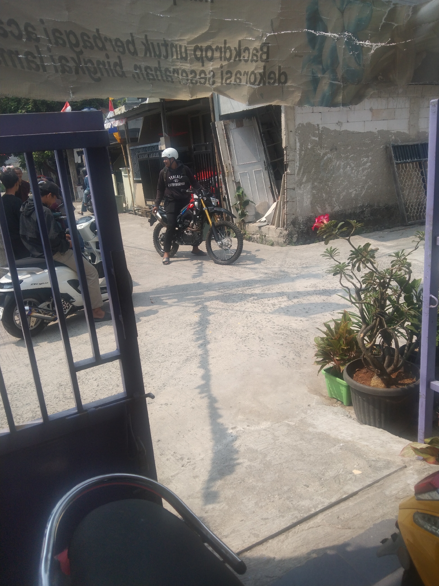 In Pantaimakmur, Indonesia, a man wearing a white helmet and a dark jacket with "TERLALU-KE-EVERYBODY" printed on the back, is seated on a red and black dirt bike, facing left. The bike has a black fender and yellow accents. To the left of the dirt bike, another person is seated on a white scooter, wearing a black jacket and beige pants, also facing left. They appear to be talking with two other individuals standing behind the scooter. In the background, a partially obscured building with grey concrete walls and a corrugated metal roof is visible, with white doors and a metal fence. The ground is paved with concrete. In the foreground, a black metal gate is partially open, revealing a view of the street scene. In the lower right corner, a yellow motorcycle is partially visible. The scene is illuminated by sunlight, casting shadows on the ground.