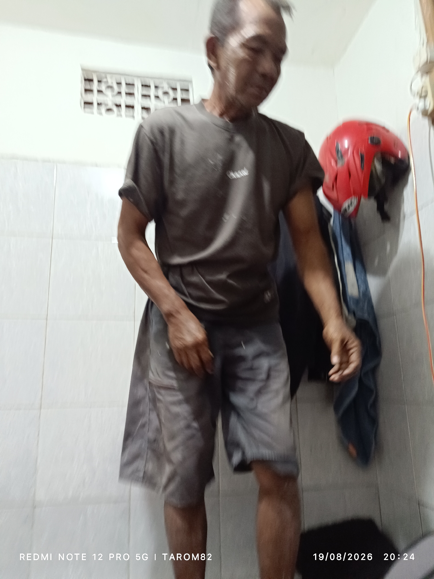 A full-body, indoor, eye-level shot shows an adult male standing in what appears to be a bathroom. The man is wearing a dark grey t-shirt and light grey shorts. He has short, grey hair and appears to be looking down and to his right. His right arm is bent at the elbow, and his right hand is held near his shorts. To the right of the man, a red helmet is hanging on the wall, next to a dark blue garment. The walls are tiled in a white, square pattern. A rectangular vent is visible on the wall behind and to the left of the man. The timestamp in the lower right corner reads "19/08/2026 20:24". The text "REDMI NOTE 12 PRO 5G | TAROM82" is visible in the lower left corner. The scene suggests a personal space, possibly in a residence in Sidodadi, Indonesia, given the timestamp's geographic indicator.