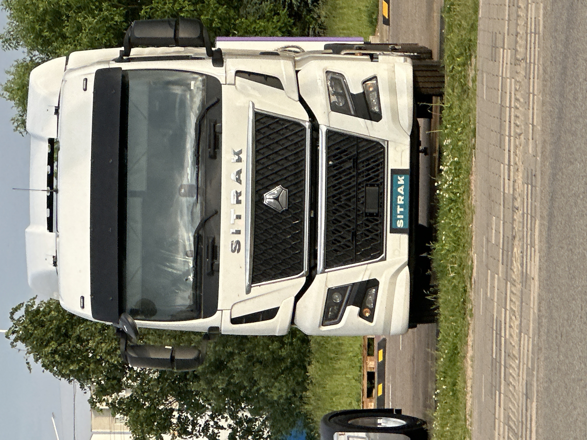 A white Sitrak heavy-duty truck is presented in a direct frontal view, parked outdoors in Бараўлянскі сельскі Савет, Belarus. The truck features a prominent black grille with a hexagonal Sitrak emblem centrally located in the upper section, and the word "SITRAK" in silver capital letters positioned above it. A lower teal-colored plate on the front bumper also displays "SITRAK" in white lettering. Integrated headlights with distinct LED daytime running lights are visible on both sides of the lower fascia. Large black side mirrors extend from the A-pillars. A black sun visor is situated above the windshield. The cab's roof exhibits white-colored structural elements and what appear to be multiple small antennas.
To the immediate left of the truck, a portion of a silver wheel with a chrome hubcap from another vehicle is visible. The ground directly in front of the truck's bumper is a narrow strip of green grass dotted with small white flowers. Below this, a grey paved surface with a textured, rectangular pattern, consistent with pedestrian paving or a sidewalk, is present, leading to a dark asphalt road surface in the foreground. In the background, behind the truck, numerous green deciduous trees are densely arrayed, partially obscuring distant light-colored buildings on the left. Wooden pallets and yellow and black striped ground barriers are visible on the ground behind the truck. The scene is illuminated by natural daylight. No individuals are present in the image.