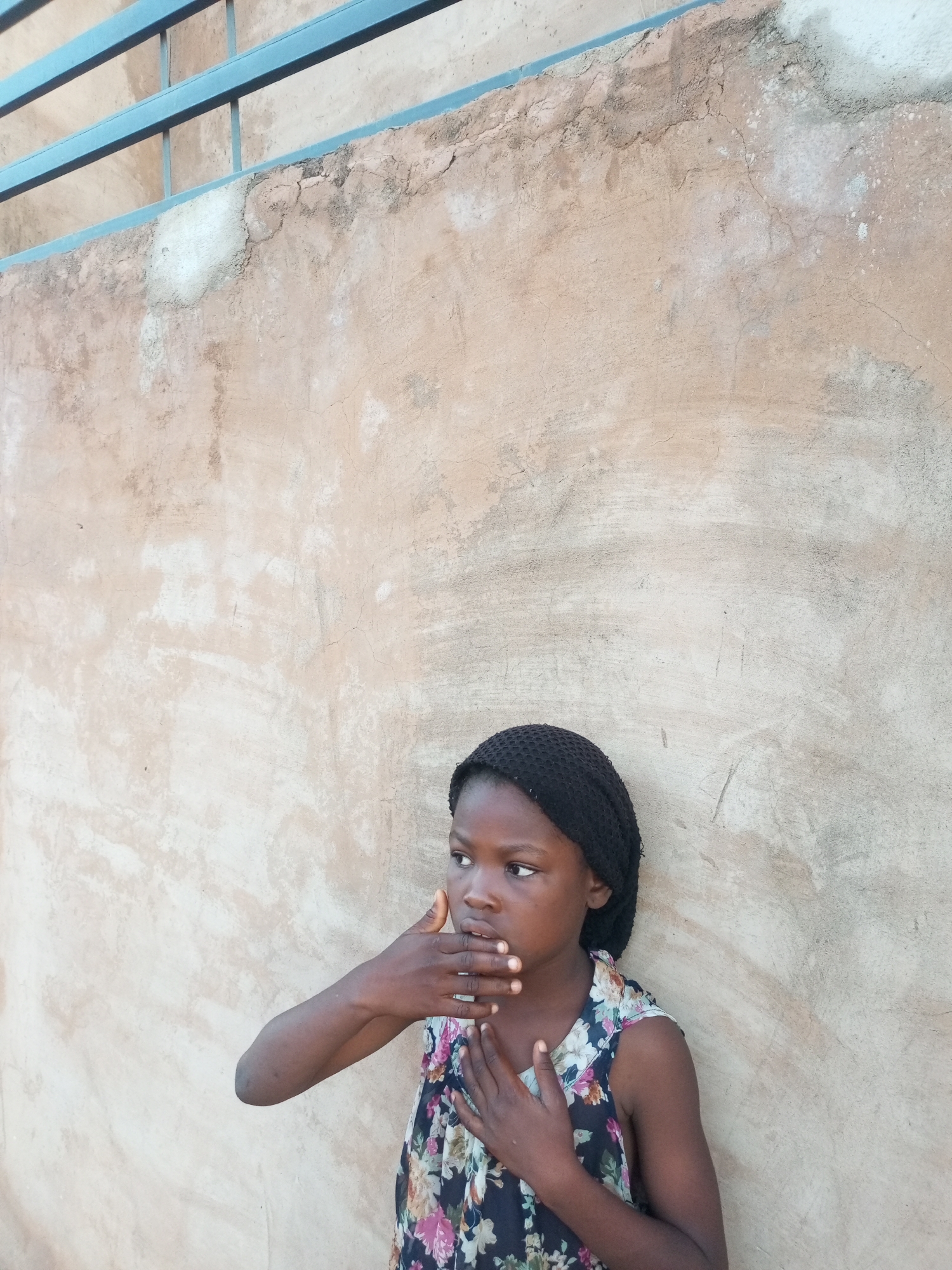 A young girl wearing a black headscarf and a floral-patterned dress stands against a textured, light-brown wall. She is looking to her left with her left hand covering her mouth and her right hand placed on her chest. Above the wall, a portion of a blue metal railing is visible against a lighter background, suggesting an outdoor setting. The wall appears weathered, with patches of lighter plaster and some cracking. The lighting suggests it is daytime. The location is identified as Tempure, Nigeria.