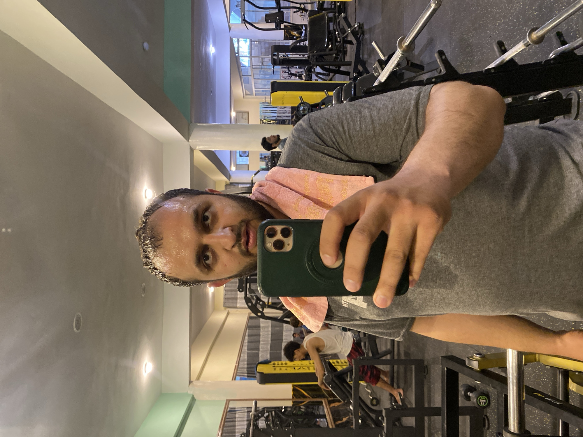 A man with a beard, sweat on his forehead and hair, is taking a selfie in a gym. He is wearing a grey t-shirt and has a pink towel draped over his shoulder. He is holding a dark green smartphone in his right hand, which is positioned in front of his face. In the background, other gym equipment and a few people can be seen. One person is sitting on a weight machine, and another is standing further back. The gym appears to be brightly lit with fluorescent lights on the ceiling. The scene suggests a casual setting within a fitness facility.
