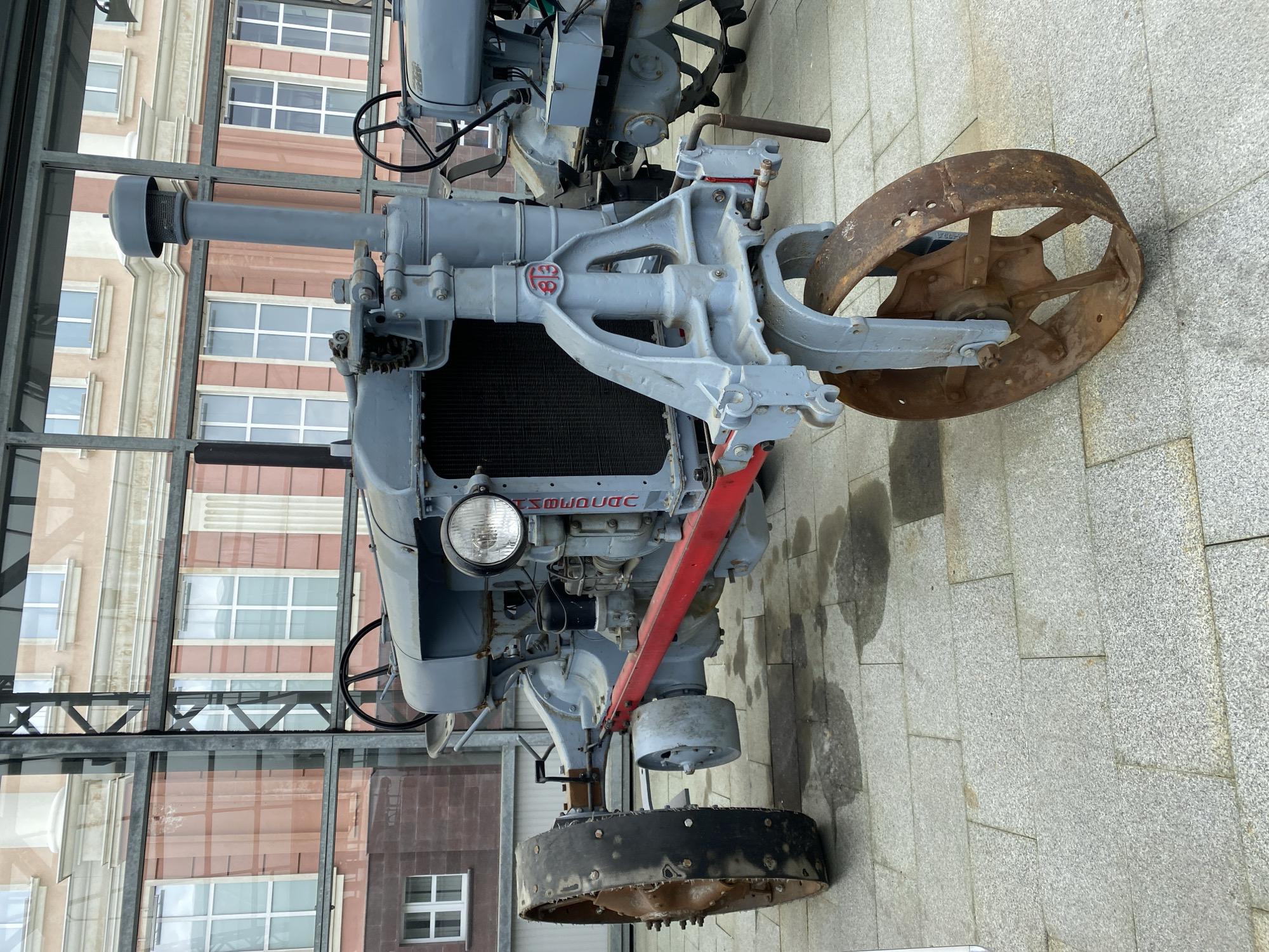 The image shows a vintage gray tractor on display at the Museum Complex of Military Equipment "Ural Military History" in Verkhnyaya Pyshma, Russia. The tractor, likely a historical exhibit, is positioned on a stone-tiled outdoor area with a glass-enclosed structure in the background, possibly part of the museum. The tractor has large, spoked metal wheels, a prominent engine area with a radiator and headlight, and a steering wheel visible. A red bar is affixed to the front chassis. The overall condition of the tractor suggests it is a restored or preserved piece of machinery. The background reveals a building with multiple windows and architectural detailing.
