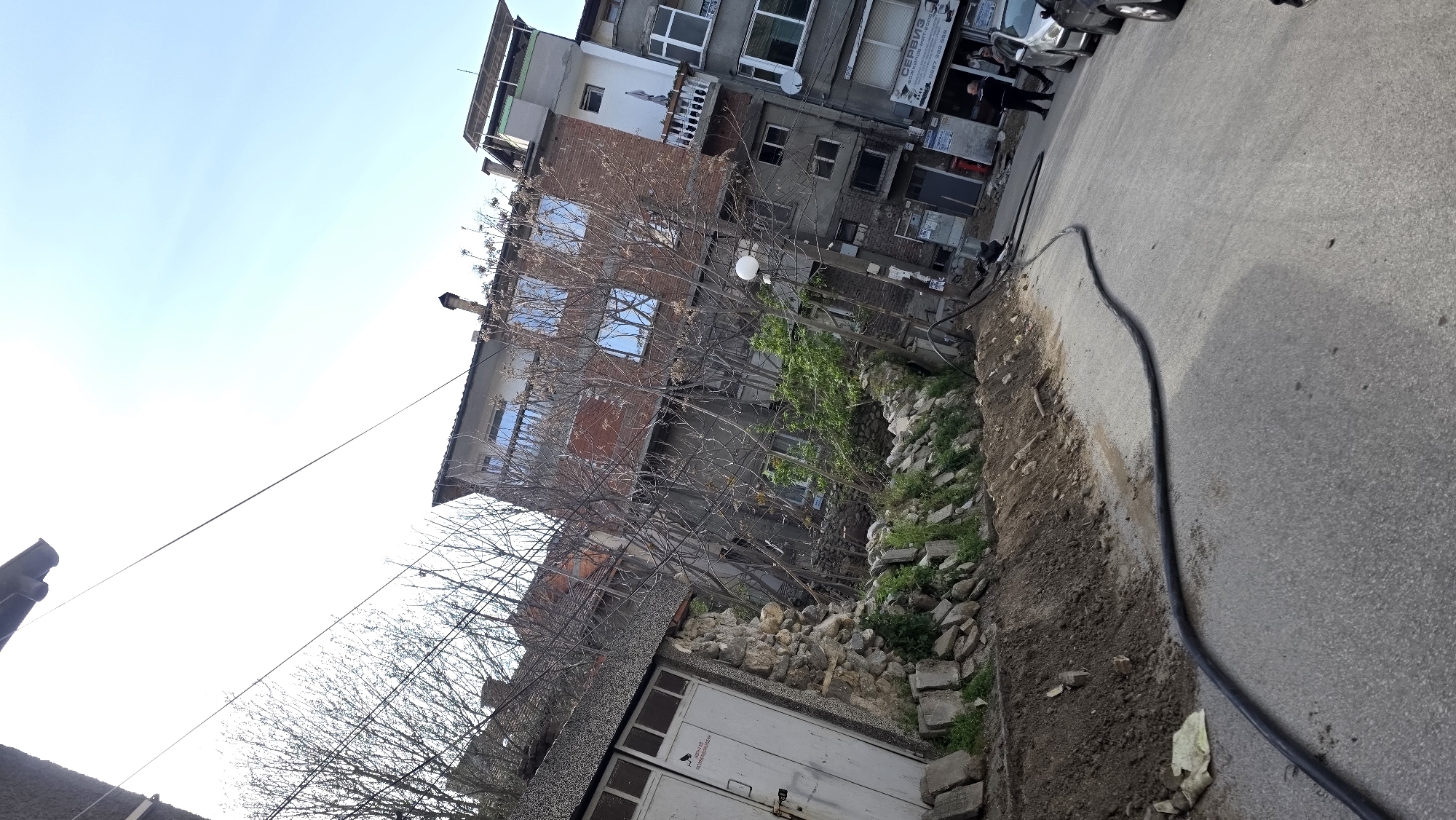 The image shows a street scene in Blagoevgrad, Bulgaria. In the background, a multi-story building with brickwork and multiple windows is visible. A narrow sidewalk borders the edge of the building. In the foreground, the street pavement is visible, with a thick black cable running along the edge of the street. A low wall constructed of stones and vegetation separates the pavement from a building with a garage door. Bare trees are between buildings. Several people are standing in front of a storefront of the building, next to parked cars. The sky is pale blue.