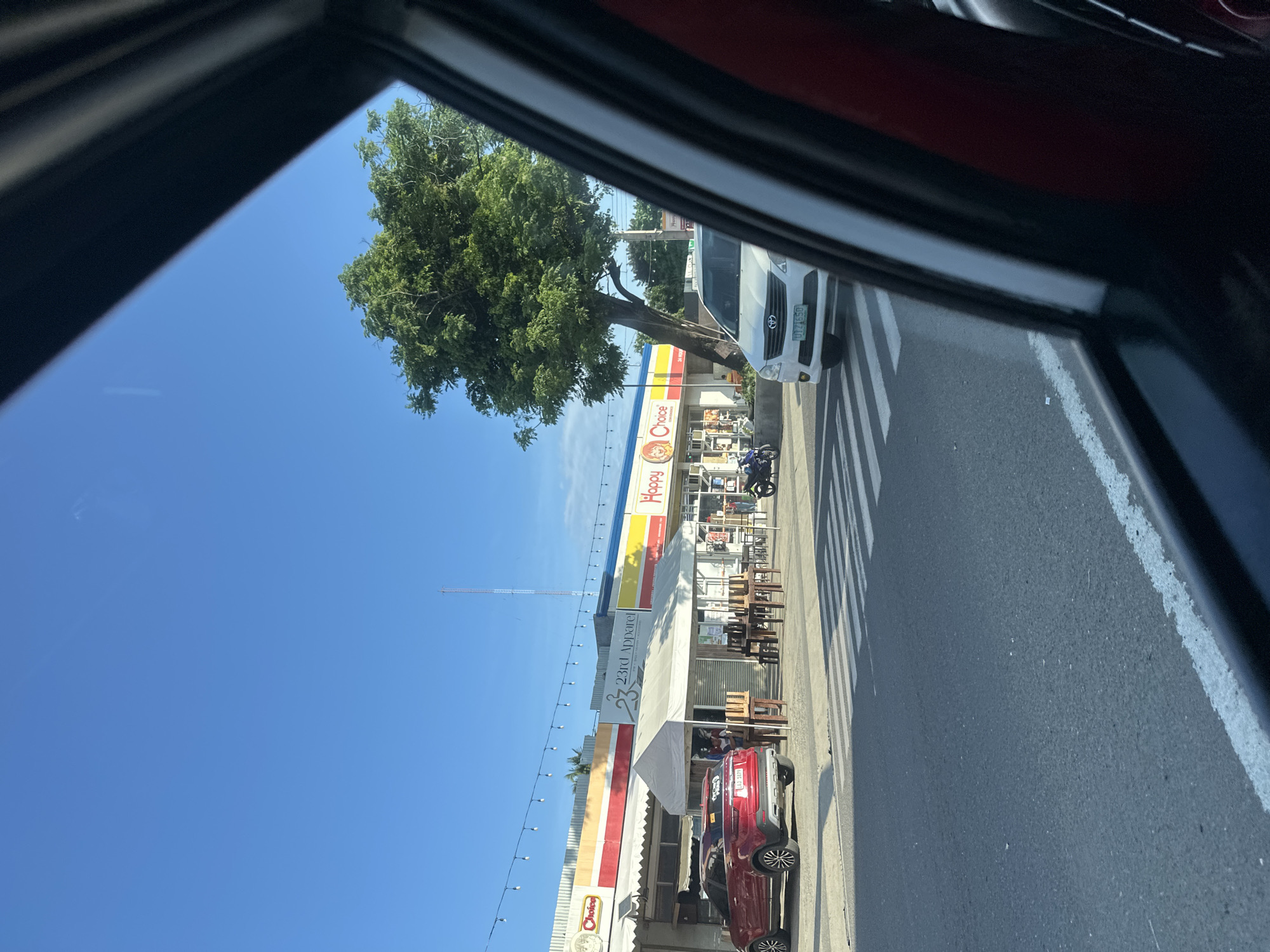The image captures an outdoor daytime street scene in Davao City, Philippines, viewed from inside a vehicle. The sky is clear blue with no visible clouds.
In the foreground, a multi-lane asphalt road is visible, featuring a white painted pedestrian crosswalk in the center and a solid white line marking the edge on the right.
Along the left side of the road, a strip of interconnected commercial buildings is present. From left to right, visible signs include "Choice" with a cartoon character logo, "23rd Apparel" with a hanger logo and the tagline "FASHION * TRENDS * ACCESSORIES", and "Happy Choice" with a smiling face logo. The buildings generally have white facades with horizontal red and yellow striped accents. String lights are strung horizontally across the front of these establishments. A white tent canopy stands in front of "23rd Apparel", under which several wooden tables are stacked, along with some chairs.
Multiple vehicles are present. A red SUV (likely a Ford Territory) is parked facing away from the viewer near the white tent; its rear license plate "LAJ 3379" is visible, along with a "CAUTION: DRIVER'S PANDEMIC" sticker on the rear window. A white Toyota Innova is positioned on the road, stopped at the crosswalk, with its front license plate "DDZ 1650" visible; it is partially obscured by the car's interior framing the right side of the image. A blue motorcycle is partially visible, parked near the "Happy Choice" establishment.
People are subtly present: one person is partially obscured near the red SUV under the tent, and another is visible near the entrance of "Happy Choice" on the far right. A large, leafy green tree stands between the commercial strip and the road, somewhat obscuring the rightmost building. The car's window frame and a portion of the interior (likely a door panel) are prominent on the right side of the image. A tall communication tower is visible in the distant background against the clear sky.