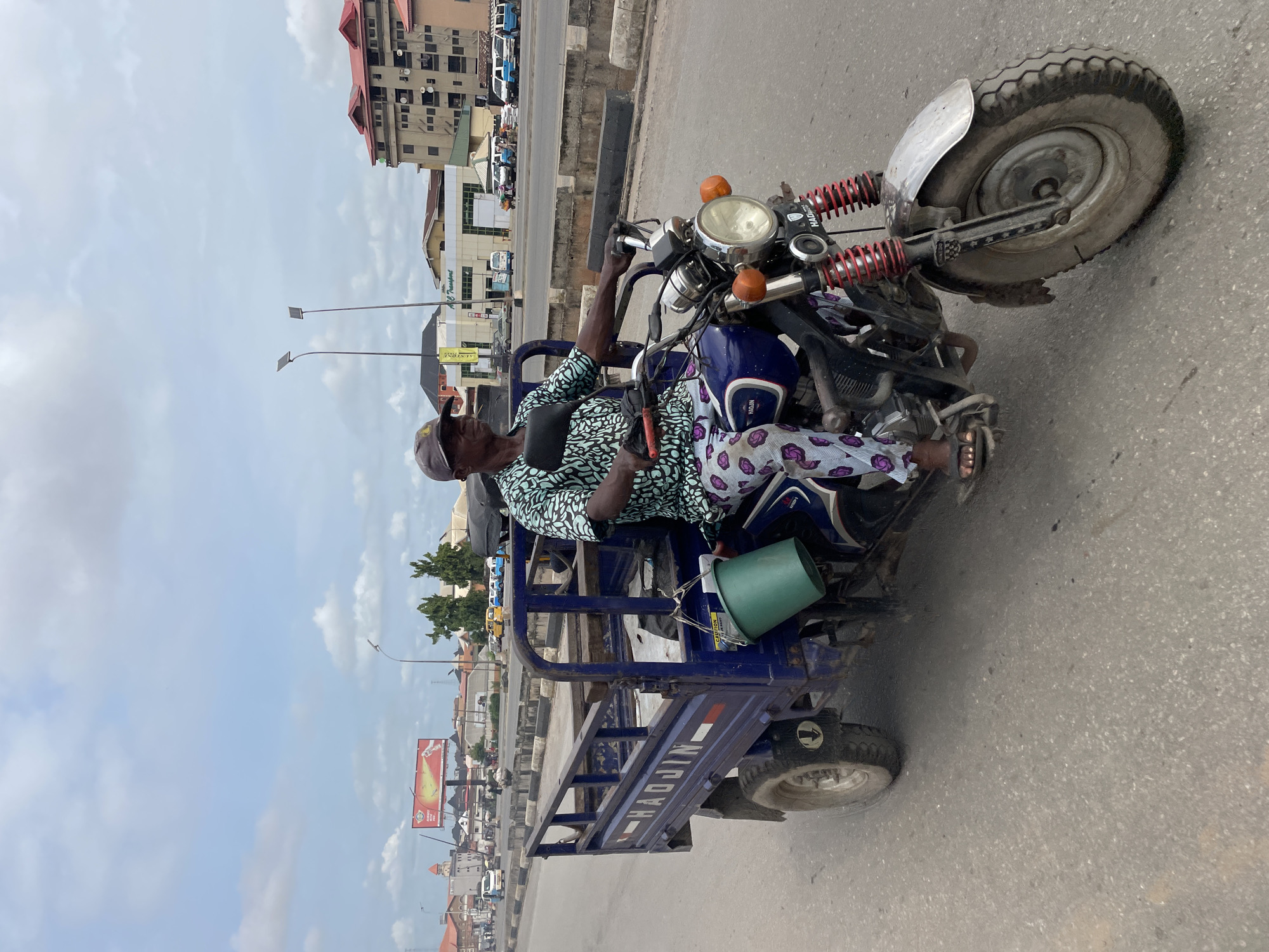 A daytime scene depicts a man operating a blue motorized tricycle on a paved road, under a partly cloudy sky. The man, wearing a light-colored cap, a light blue/green and white patterned shirt, and purple and white patterned trousers, is seated in the driver's position, looking forward. A small, dark pouch is visible across his chest.
The tricycle is a three-wheeled vehicle featuring a motorcycle front section with a round headlight, orange indicator lights, and prominent red coil springs on the front suspension. Attached to the rear is a sturdy, blue cargo bed. A green plastic bucket is tied to the side rail of the cargo bed. The brand name "HAOJIN" is clearly visible in white letters on the side panel of the cargo section. The vehicle's wheels are robust, suggesting it's designed for varied conditions.
The background reveals an urban or semi-urban environment with multiple buildings lining the roadside. These include multi-story structures with different roof types, some with flat roofs and others with sloped, red roofs. Faintly visible on one building's facade is "M.L Transport". The road appears to be a major thoroughfare, with other vehicles and traffic visible in the distance. Streetlights, some equipped with solar panels, are spaced along the road. A large yellow and red billboard is also seen further down the street. The weather is clear and bright, indicating good visibility.