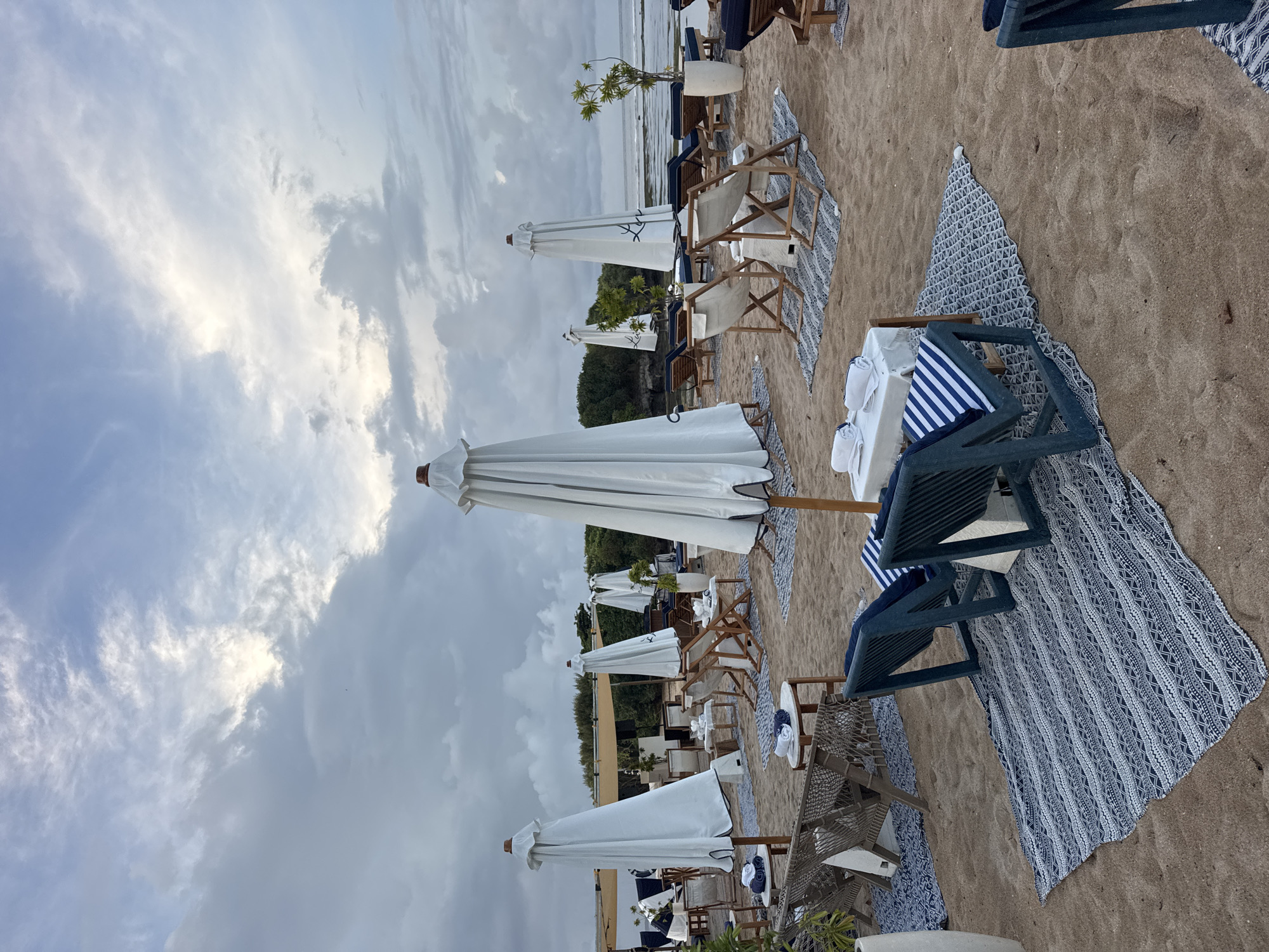 The image depicts a beach scene in Kuta Selatan, Indonesia, with rows of beach chairs and white sun umbrellas positioned on the sand. The beach furniture is set up in a neat arrangement, each set accompanied by patterned rugs and small tables. The sky is overcast with heavy clouds, and the ocean can be seen in the background. There are no people visible in the immediate scene, but the setting suggests a relaxed, tourist-oriented environment.