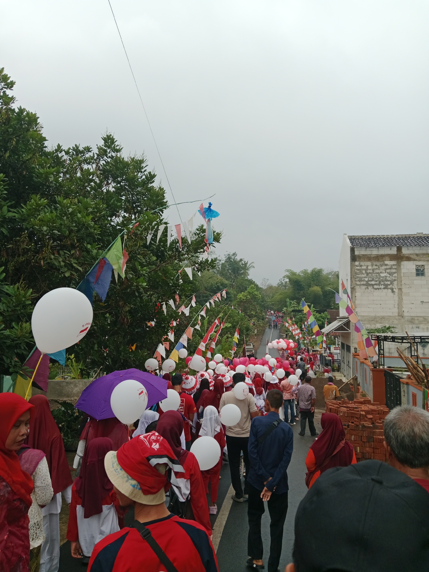 The image depicts a street procession in Bojongmengger, Indonesia, likely for a national celebration, evidenced by the red and white decorations and Indonesian flags. A large group of people, many wearing red and white attire, are walking down a paved road lined with trees and houses. Some participants are carrying white balloons, and a purple umbrella is visible among the crowd. The sky is overcast, suggesting a cloudy or rainy day. Construction materials, including stacks of bricks, are present along the right side of the street, indicating ongoing development or renovation in the area. The overall scene suggests a community event or parade.