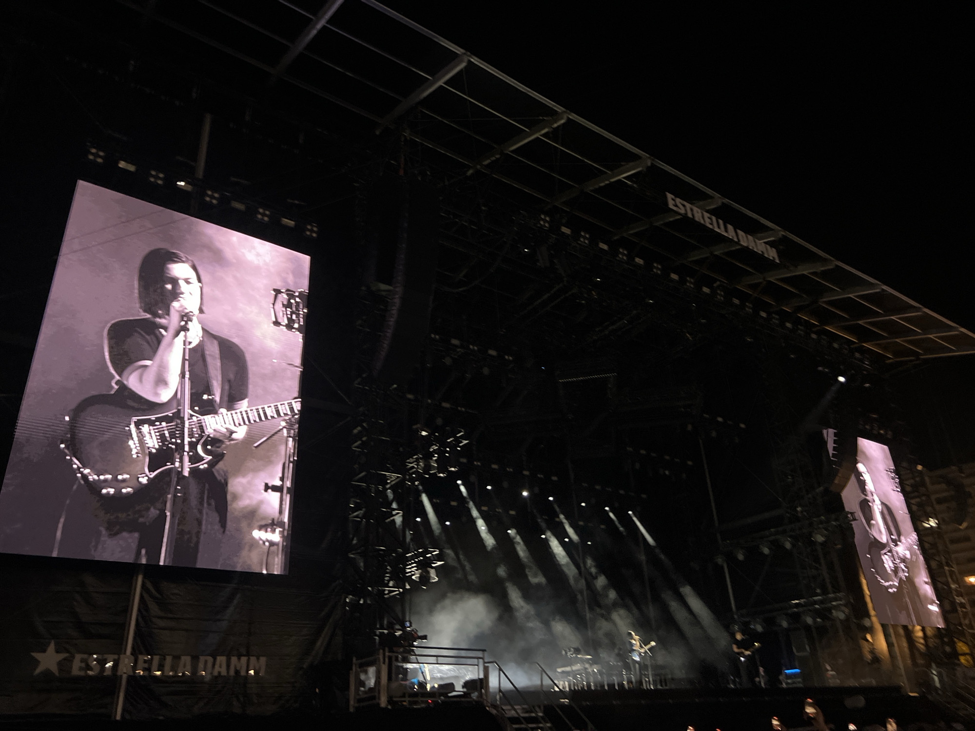 The image captures a concert at night in Porto, Portugal, likely during the Primavera Sound festival, as evidenced by the "Estrella Damm" branding visible on banners and the stage structure. The scene is dominated by a large stage featuring prominent video screens displaying a black and white image of a musician playing a guitar. The stage is equipped with elaborate lighting rigs and speakers. Smoke or fog machines are active, creating atmospheric plumes illuminated by spotlights. In the background, a crowd of concert-goers can be seen, their hands raised. The overall atmosphere is that of a large-scale outdoor music event.