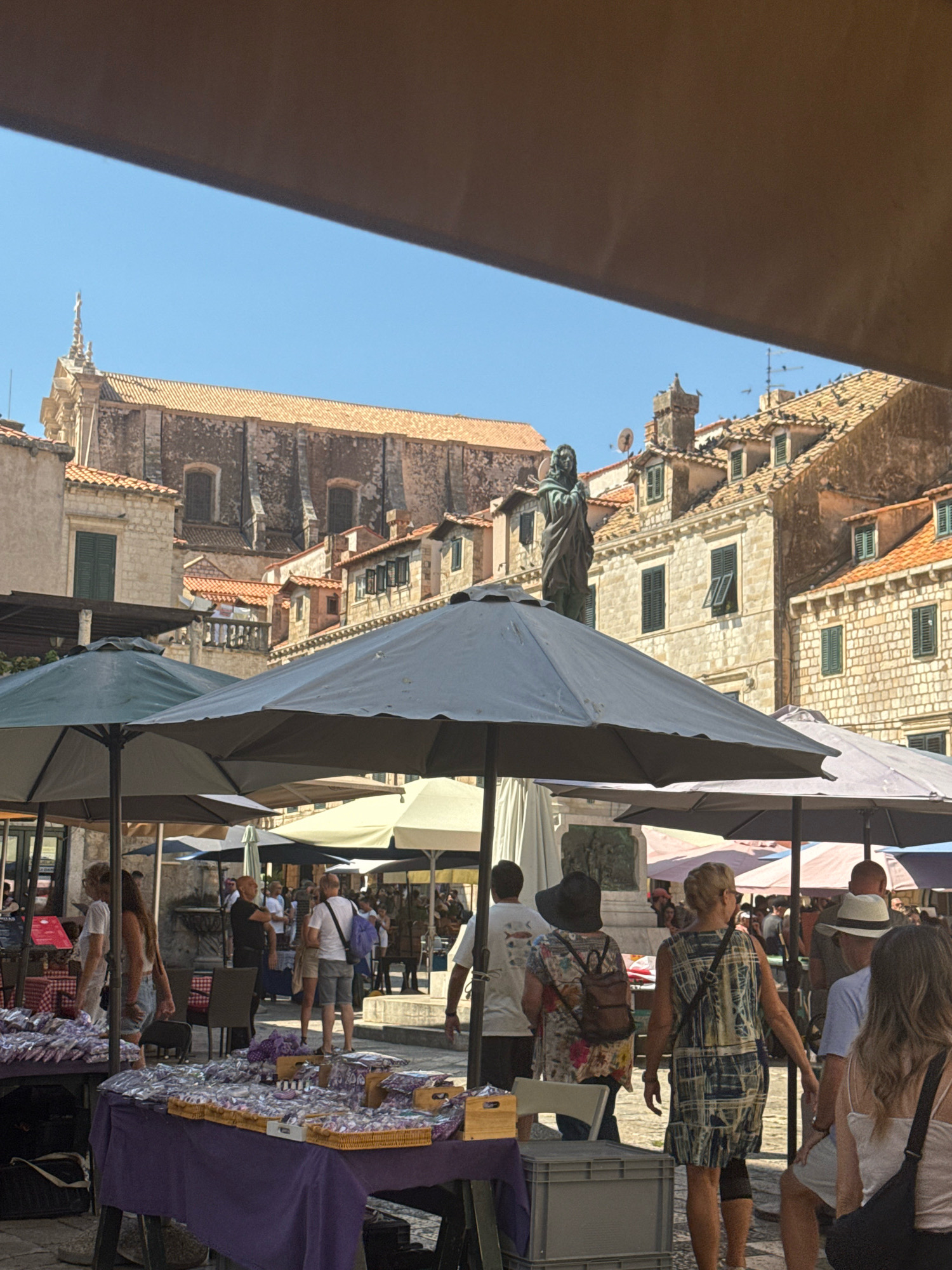 This image depicts a bustling market scene within the city of Dubrovnik, Croatia, likely in the historic Old Town. In the foreground, several large, open umbrellas provide shade over market stalls laden with goods. The stalls are draped with purple tablecloths and display numerous small packages, some appearing to be lavender products. Several people are visible browsing the stalls and walking through the market. In the middle ground, a bronze statue stands atop a pedestal, appearing to be a historical or religious figure. Behind the market and statue, traditional stone buildings with terracotta roofs characteristic of Dubrovnik architecture are visible. The clear blue sky indicates fair weather. The overall impression is one of a vibrant and active outdoor market in a historic European city.