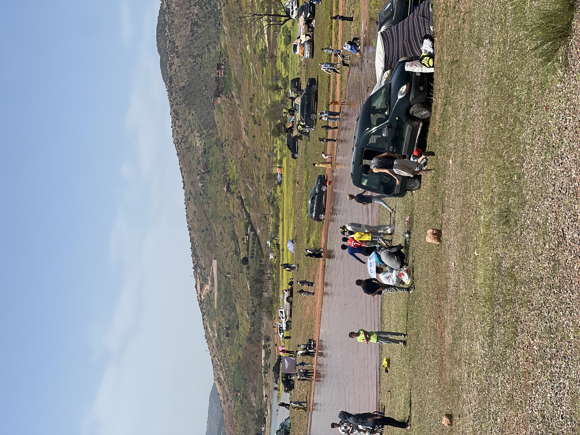The image depicts a scene in Bin El Ouidane, Morocco, showing a large, open area possibly near a lake or river. The ground is a mix of dirt and grass, appearing wet or flooded in certain areas. Several people are present, scattered around, some gathered in groups, possibly interacting or setting up temporary camps. There are cars parked along the periphery of the open area, some with items placed on top or around them, suggesting a recreational or gathering event. The environment includes a large hill or mountain in the background, providing a natural backdrop. The sky is visible, appearing clear and bright, indicating it is daytime. The overall impression is of an informal gathering or event taking place in a natural setting, possibly after rainfall, near the lake of Bin El Ouidane.