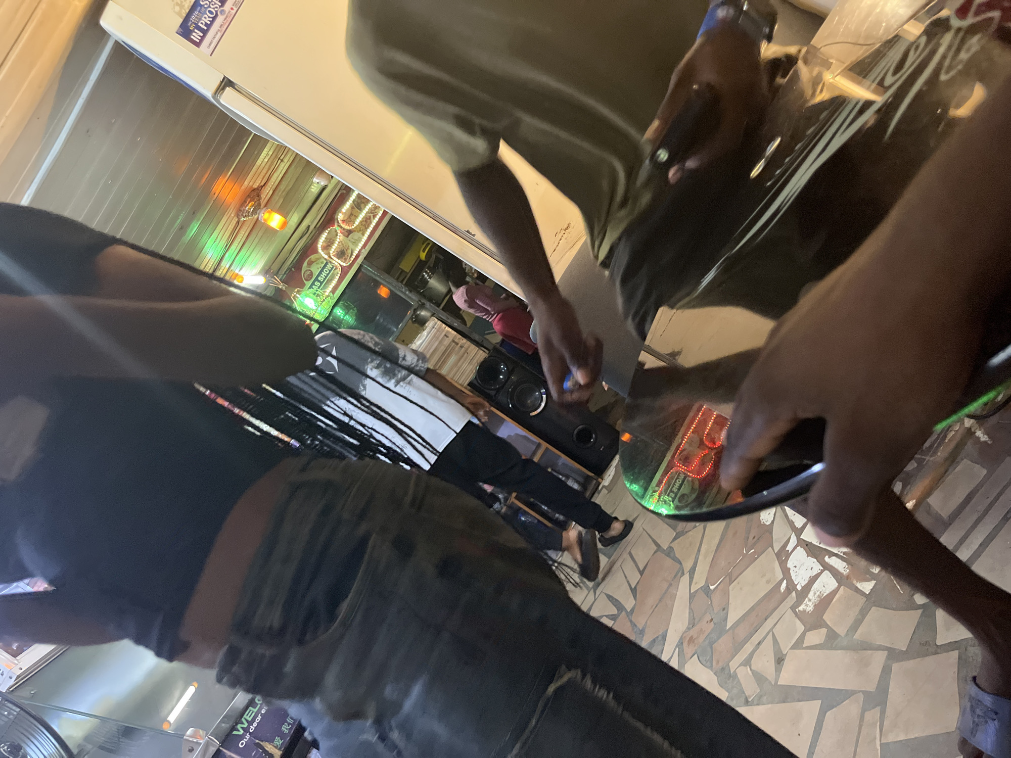 A medium shot captures a dimly lit interior scene, possibly a shop or lounge, with a focus on the lower bodies of several individuals and various objects. The foreground features the hands of a person holding a dark object, possibly a phone or remote, with a reflective surface, perhaps a table, below. To the left, a person is wearing blue flip-flops and denim jeans. Another individual in the background is seen wearing a light-colored t-shirt and dark pants, with their feet bare. To the left of this person, a tall, black speaker is visible. Further in the background, a woman in a pink headscarf and red skirt is standing near shelves stocked with items. The environment is characterized by a patterned tiled floor and a metallic ceiling with colorful lights, including green and orange. A white refrigerator is positioned on the right side of the frame, displaying stickers. The lighting is low, with some areas illuminated by the colored lights and what appears to be a refrigerator light.